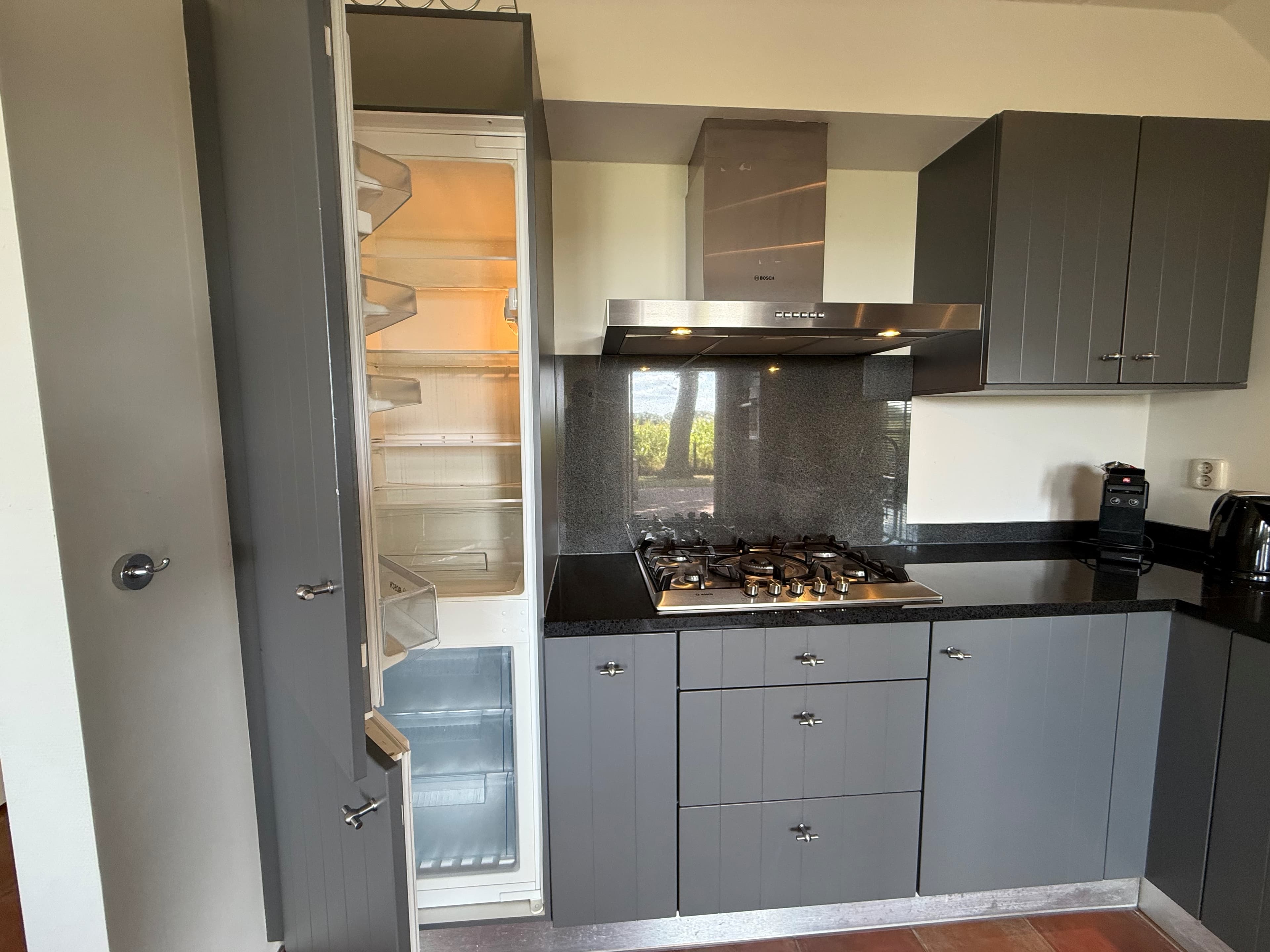 Keuken met grijze kastjes, gasfornuis en een open koelkast met transparante planken.
