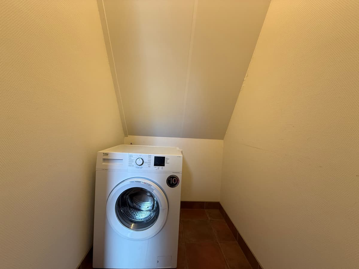 Waskamer met een wasmachine onder een schuin plafond en witte wanden in een recreatiewoning.