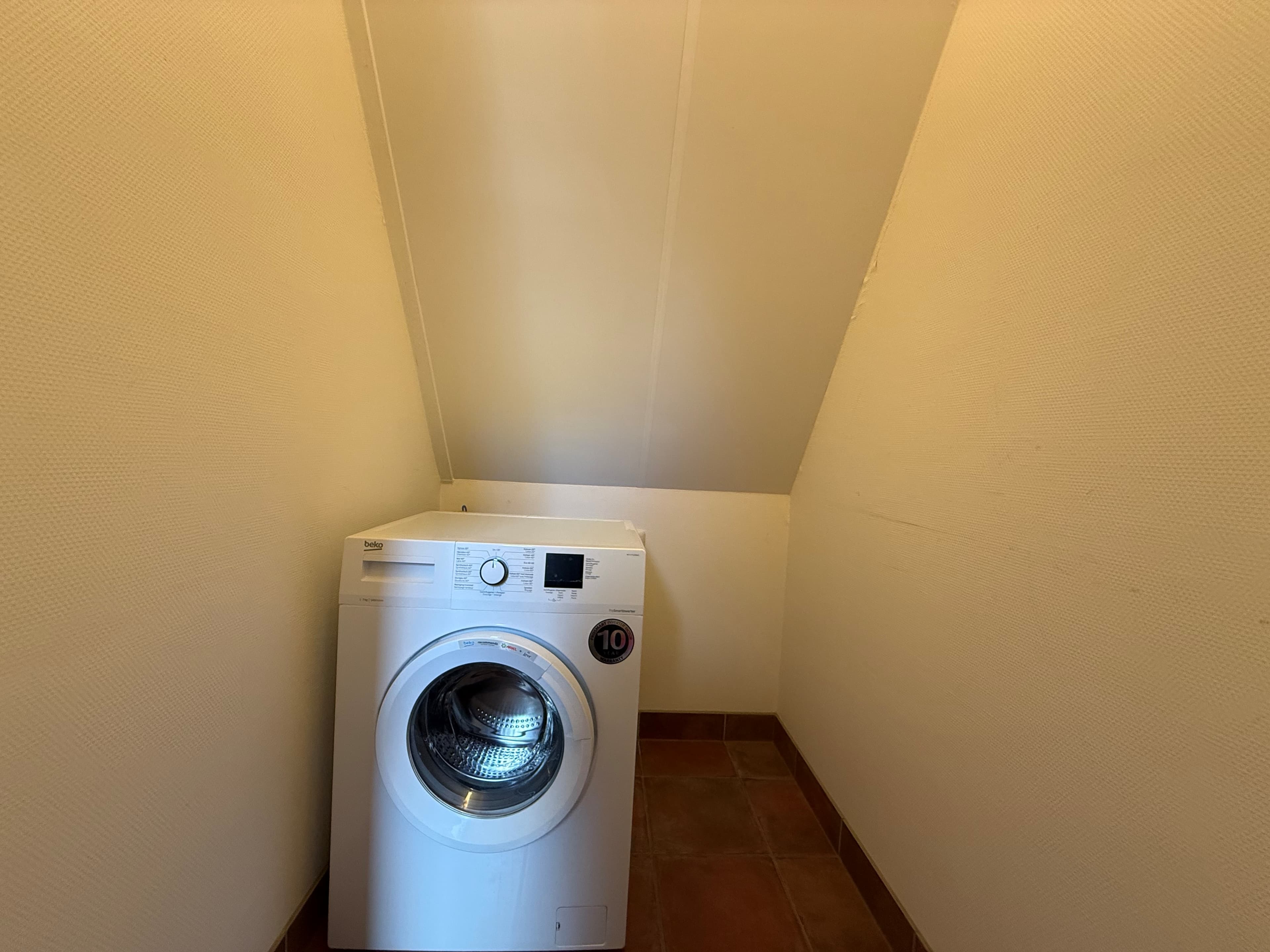 Waskamer met een wasmachine onder een schuin plafond en witte wanden in een recreatiewoning.
