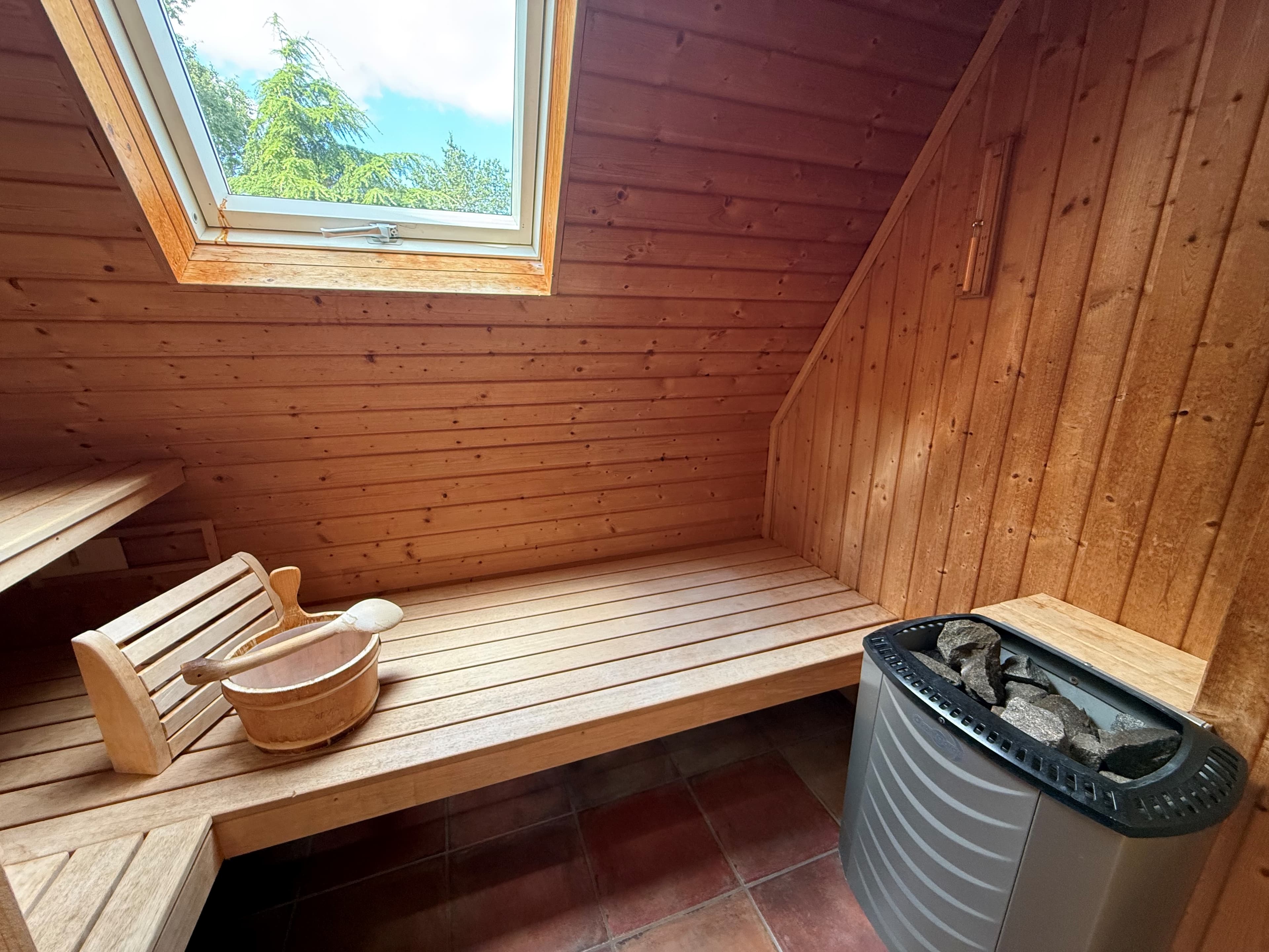 Sauna met houten wanden, banken en een venster, inclusief een stenen kachel en een houten emmer.