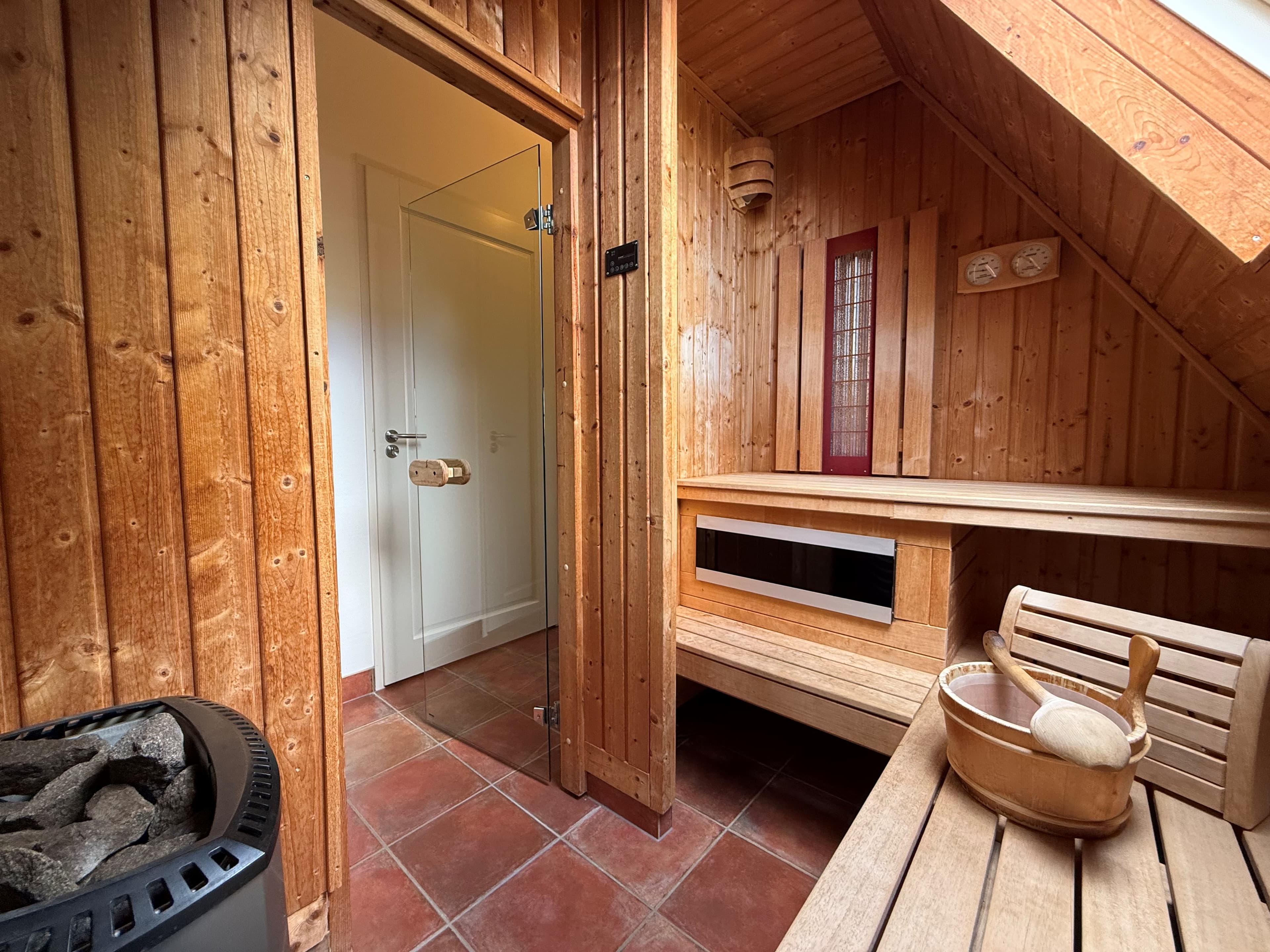 Binnenzicht van een sauna met houten wanden, een glazen deur, een zitbank en een haard.