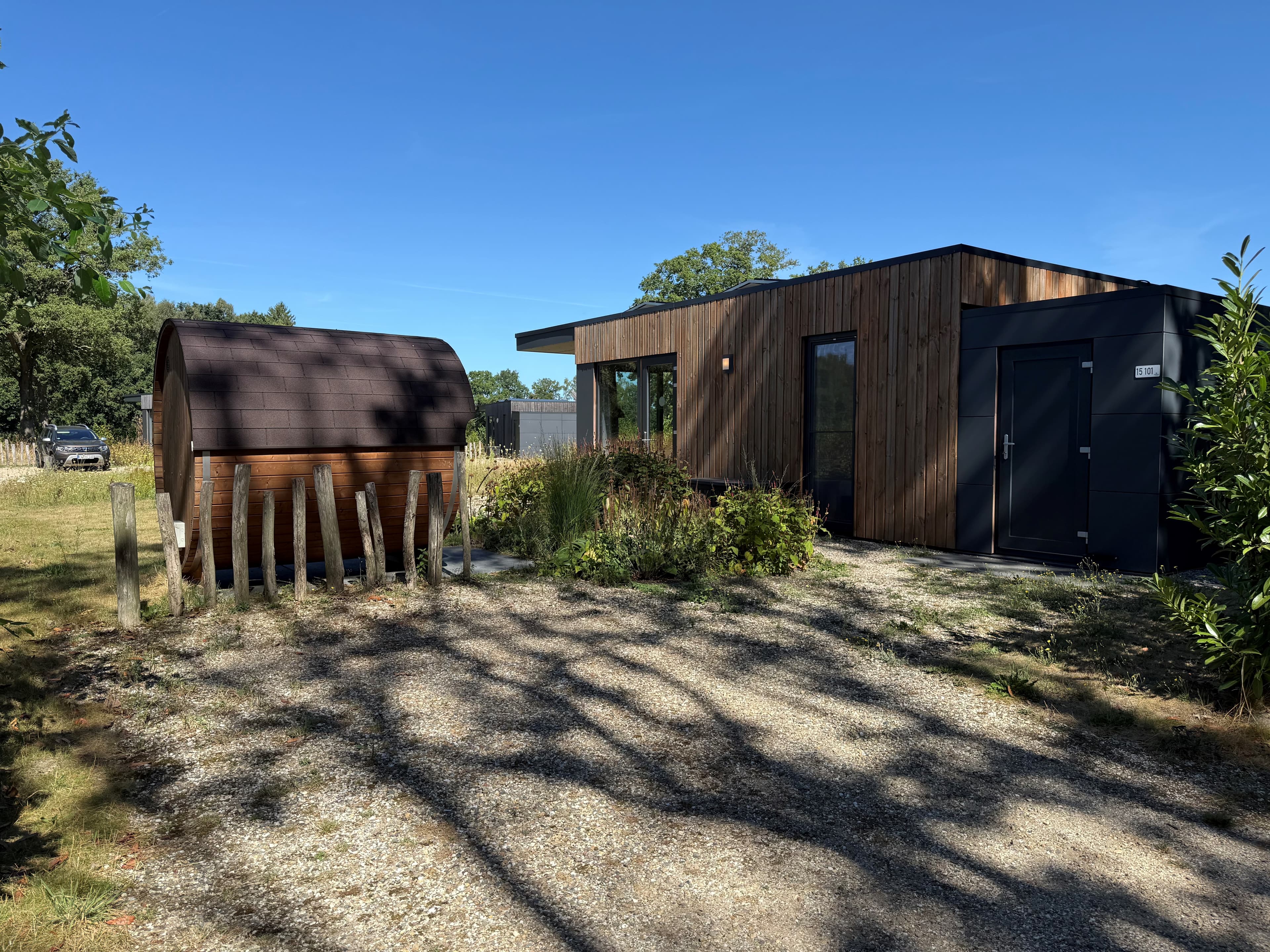Moderne vakantiewoning met houten gevel en een houten sauna aan de zijkant, omgeven door groen en een grindpad