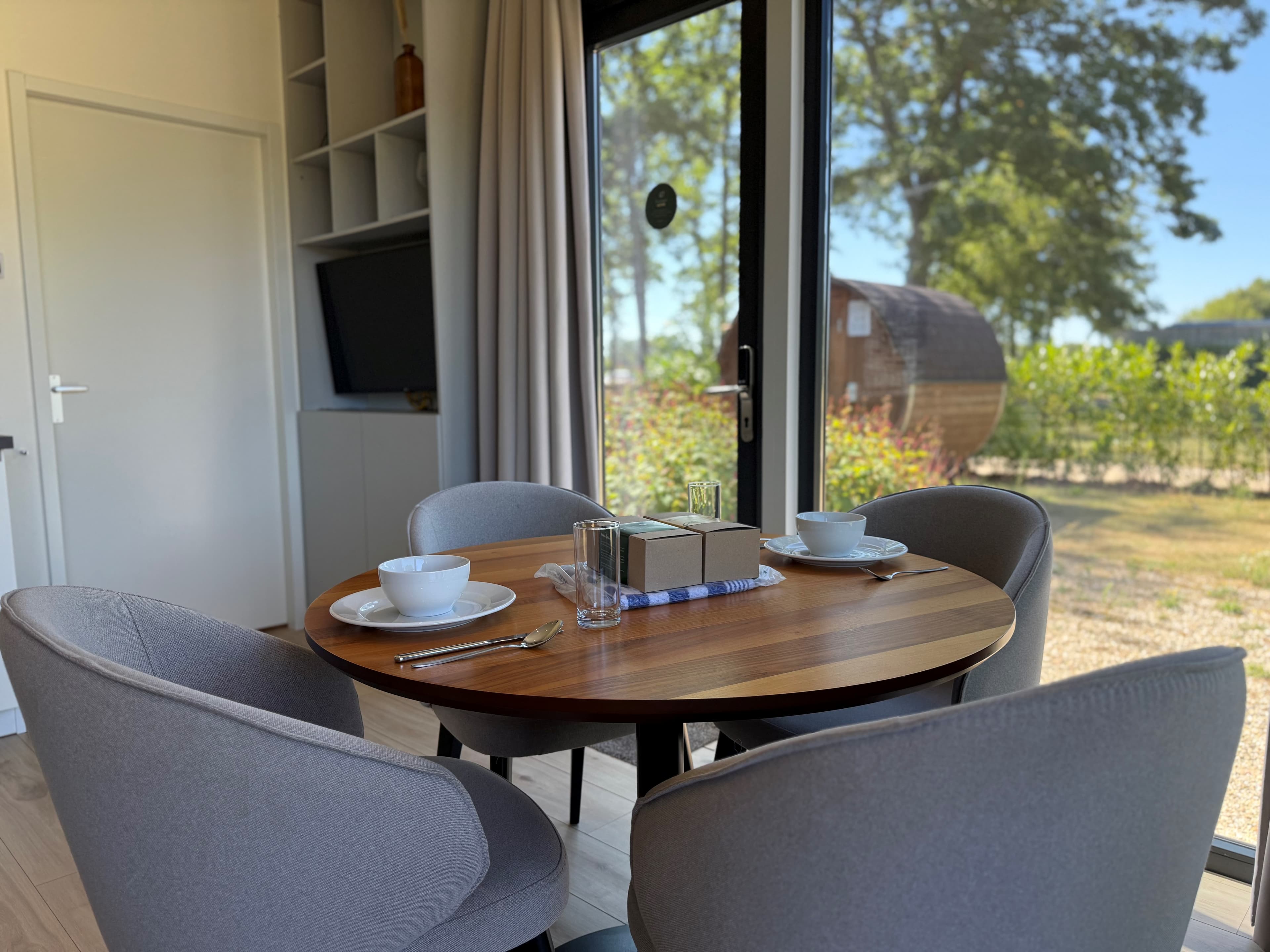 Eetgedeelte met ronde houten tafel en vier grijze stoelen, grote ramen met uitzicht op de tuin en natuur buiten.
