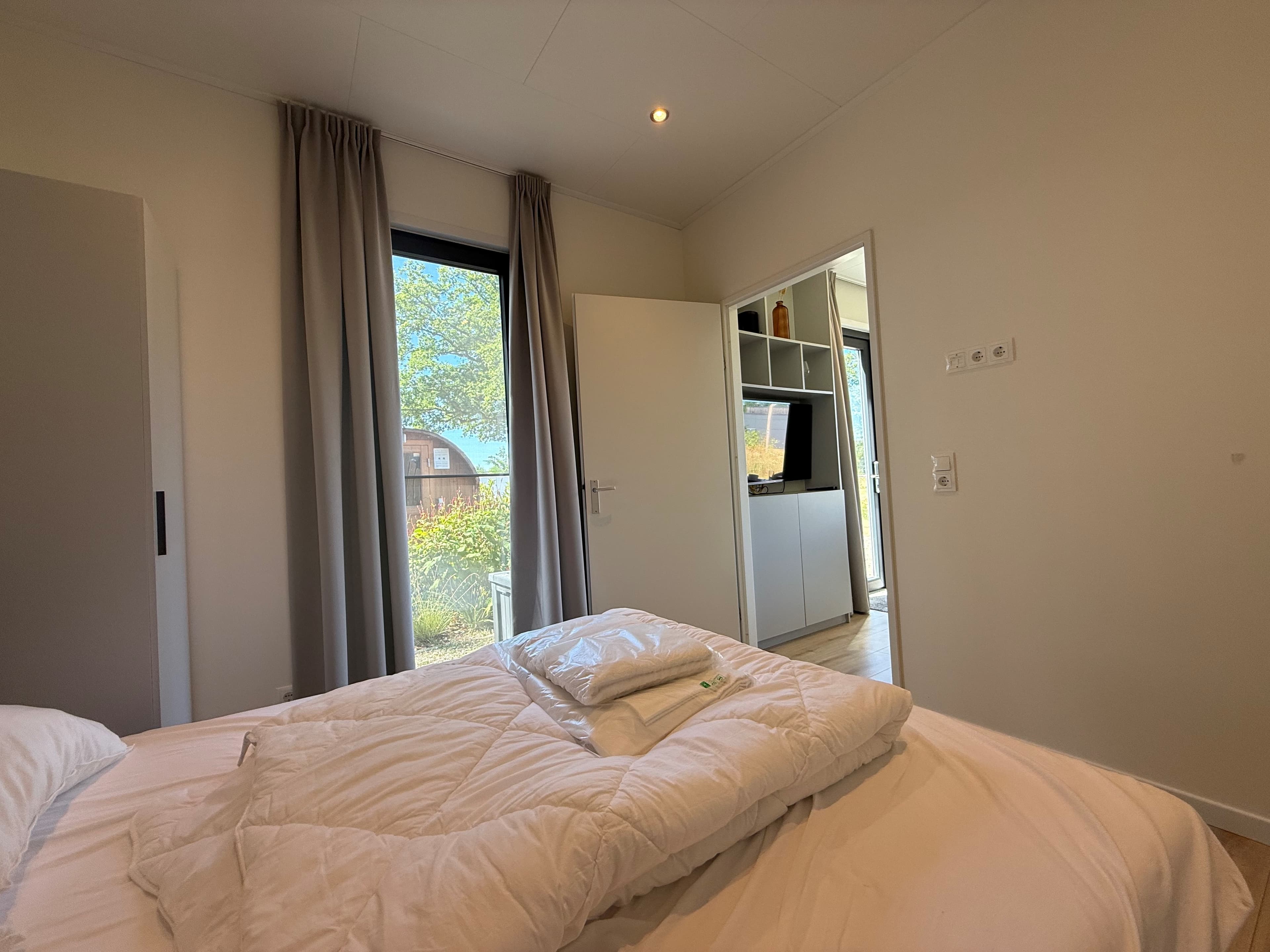 Slaapkamer met een bed en kasten, grote ramen met uitzicht op de tuin en een deur naar een andere ruimte.
