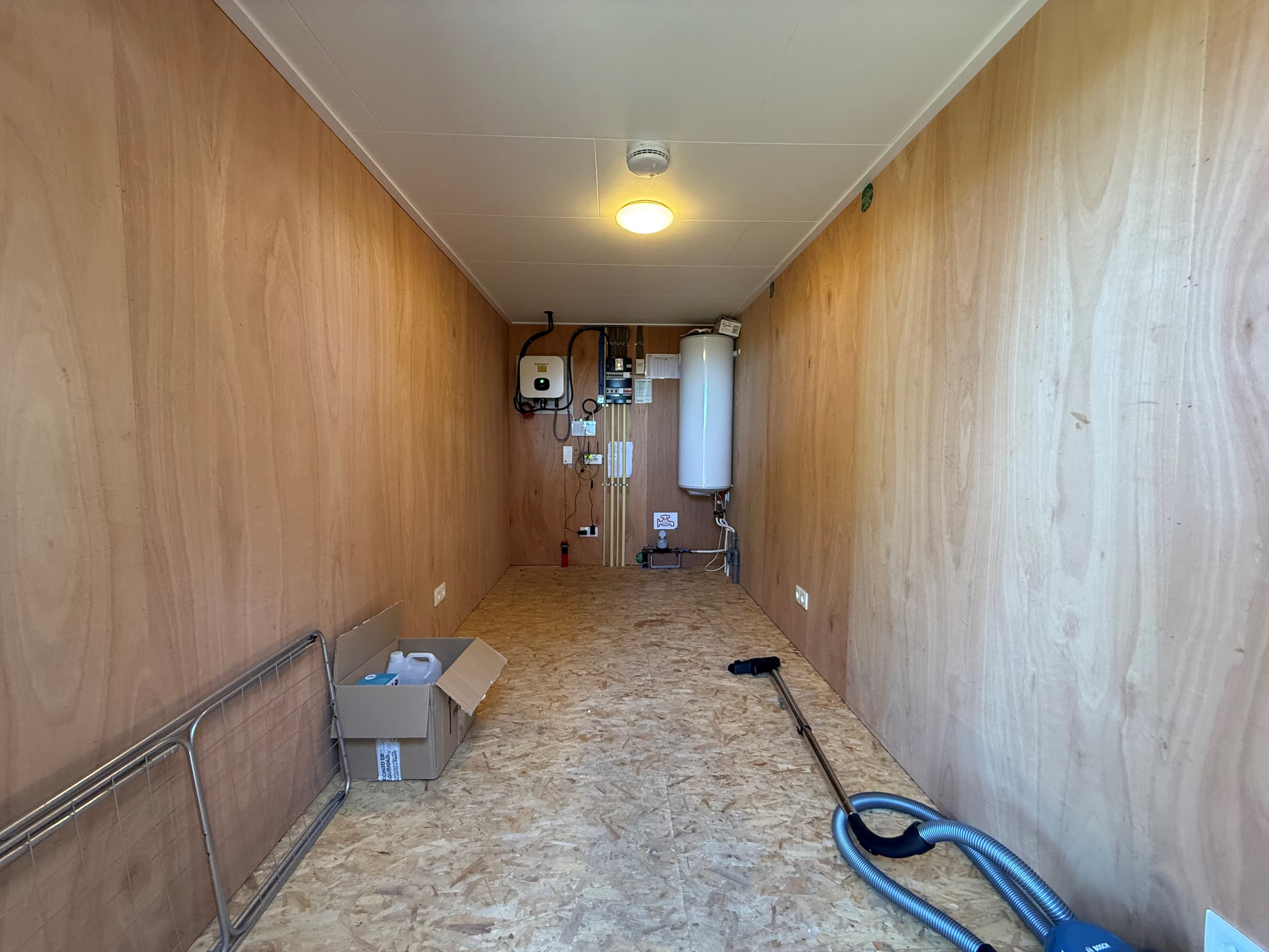 Lege ruimte met houten wanden, een vloer van OSB-platen en enkele installaties aan de muur, inclusief boiler en leidingen