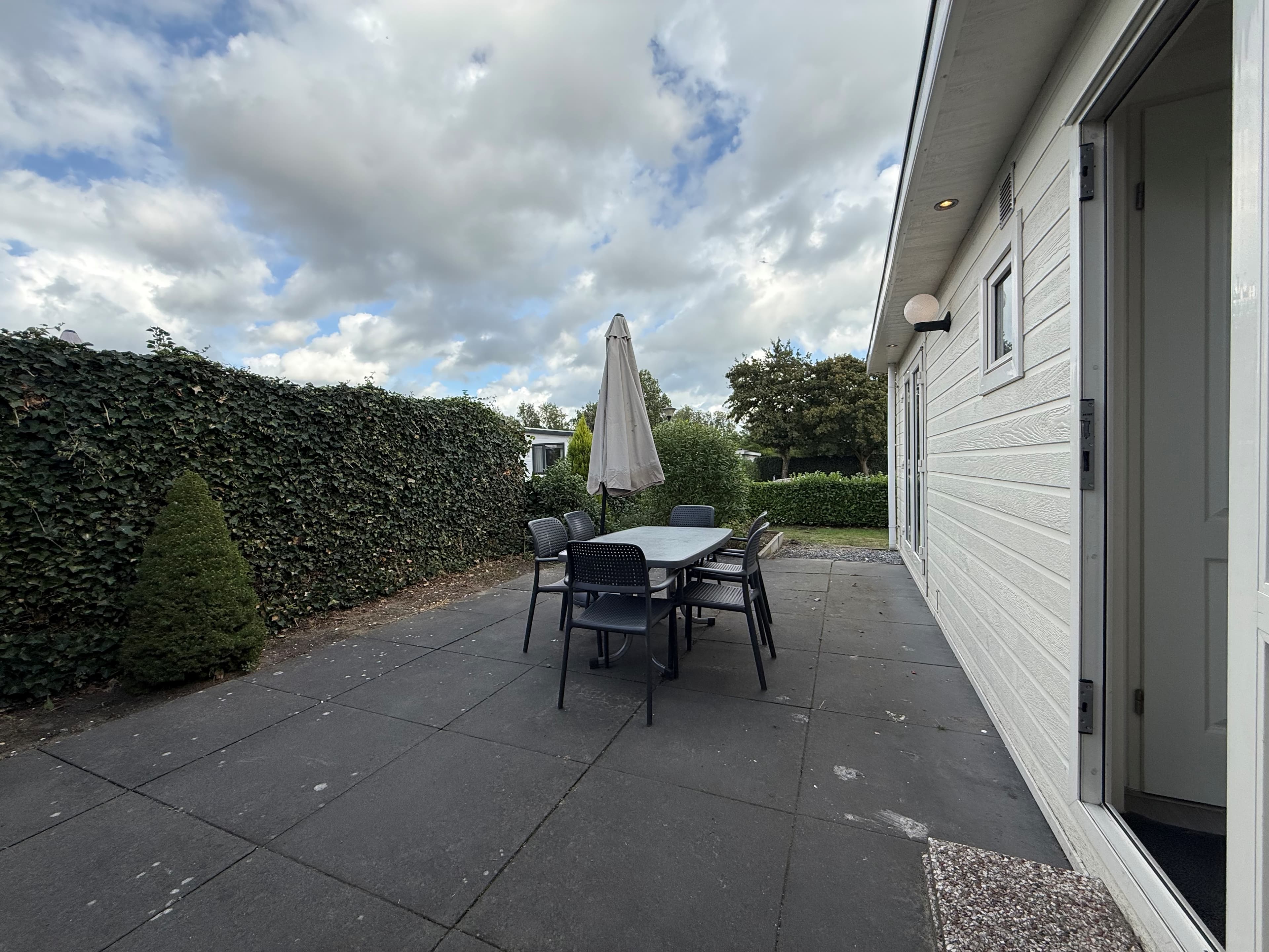 Terras met eettafel en stoelen bij een recreatiewoning, omringd door groene hagen en een blauwe lucht met wolken