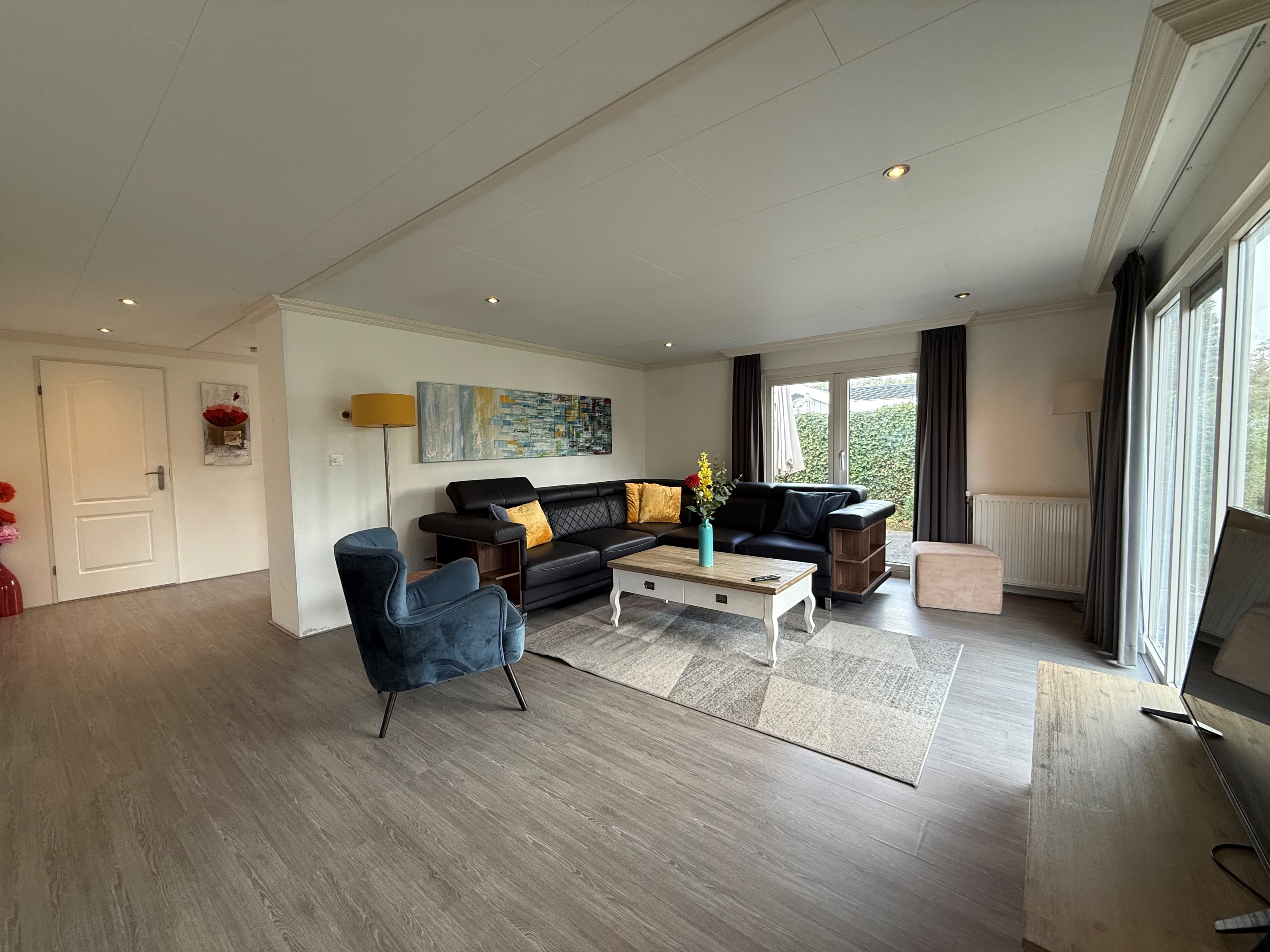 Lichte woonkamer met houten vloer, zithoek met bank en stoelen, grote ramen en decoratieve elementen op de wanden