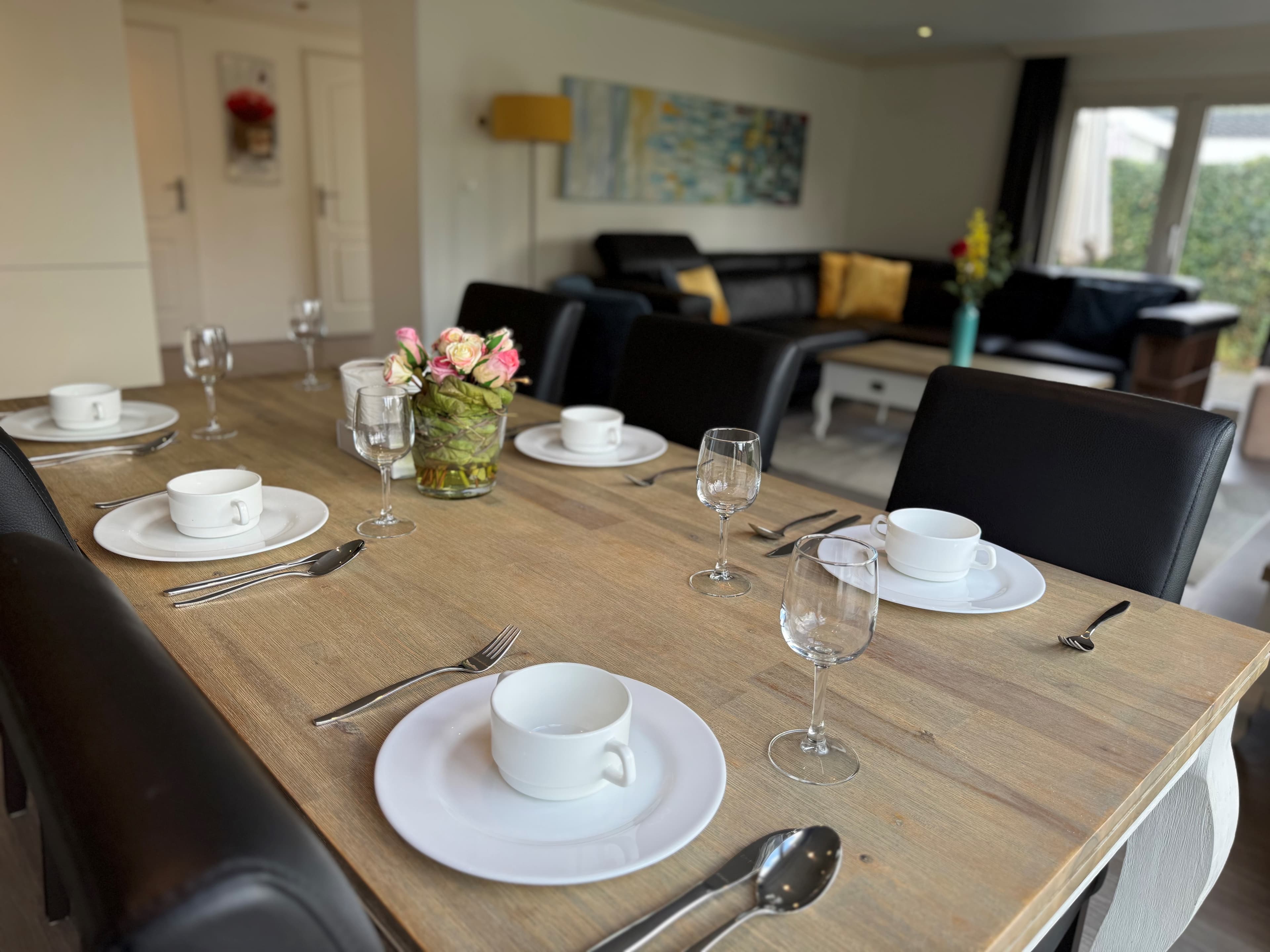 Eettafel met witte borden en glazen, bloemenvaas op tafel, zithoek op achtergrond met bank en kleurrijke schilderij.