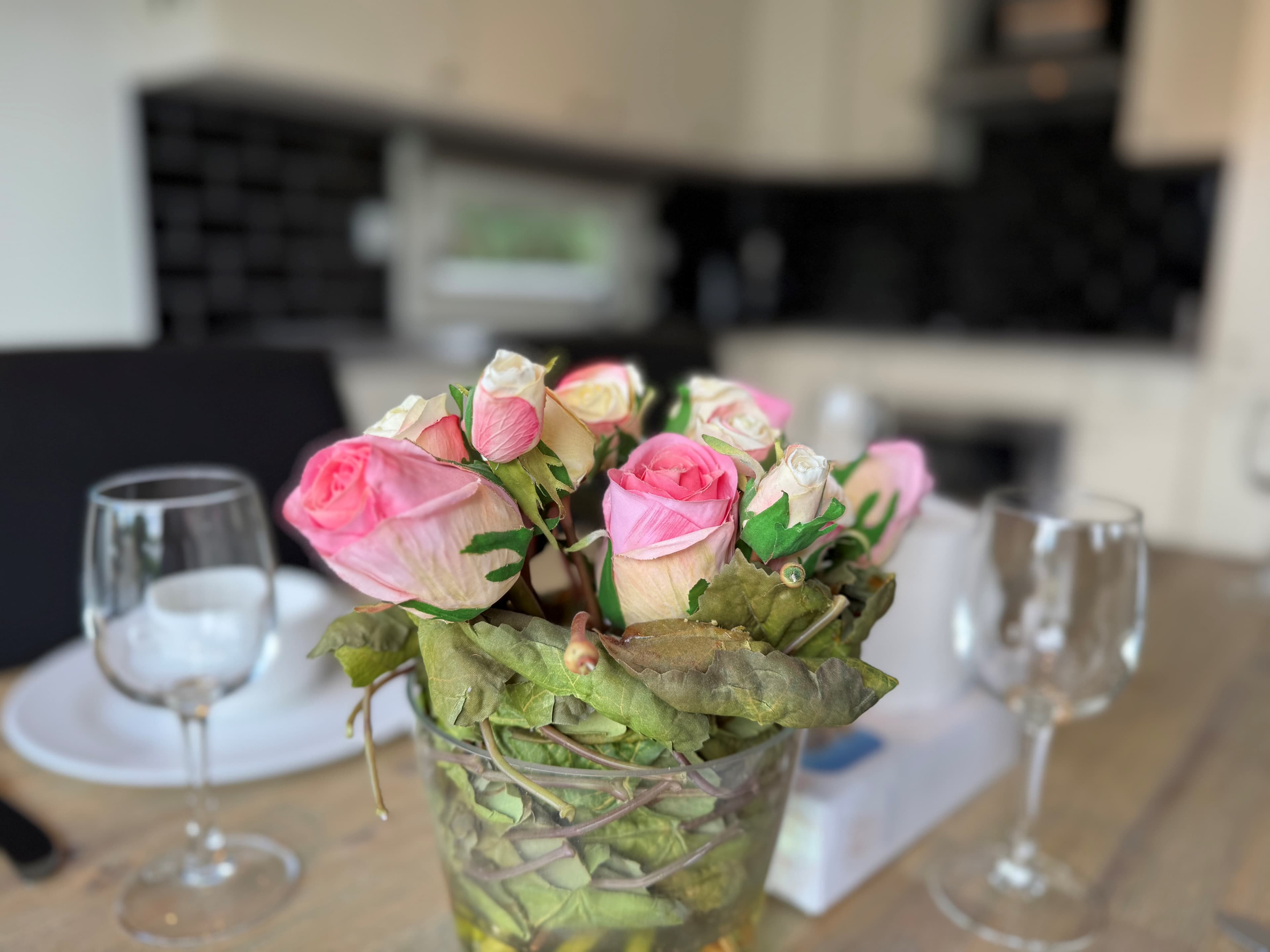 Bloemstuk met roze rozen in een glazen vaas op een houten tafel, met een servies en glazen op de achtergrond