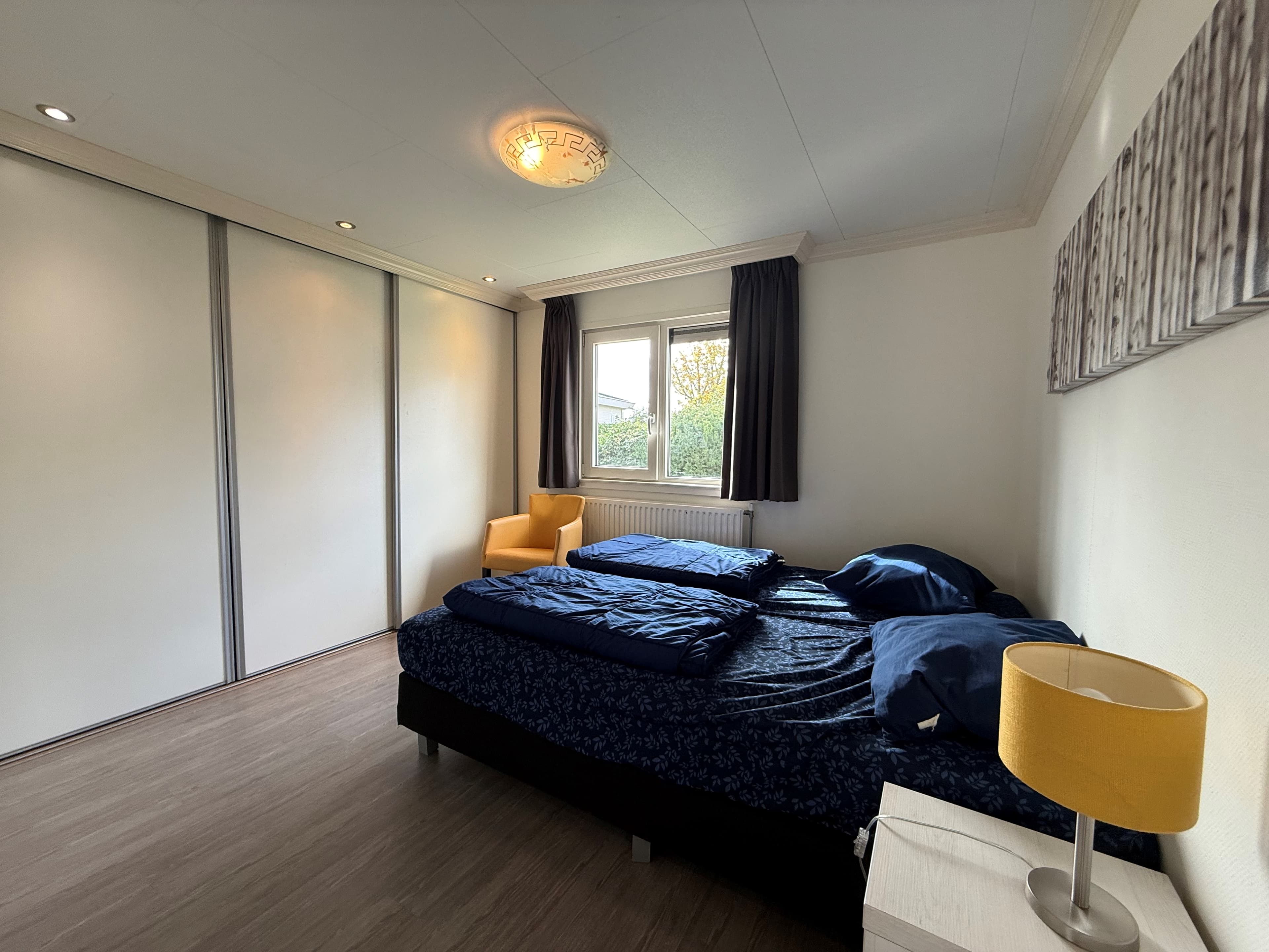 Slaapkamer met twee eenpersoonsbedden, een gele fauteuil, een nachtkastje en een kledingkast met schuifdeuren.