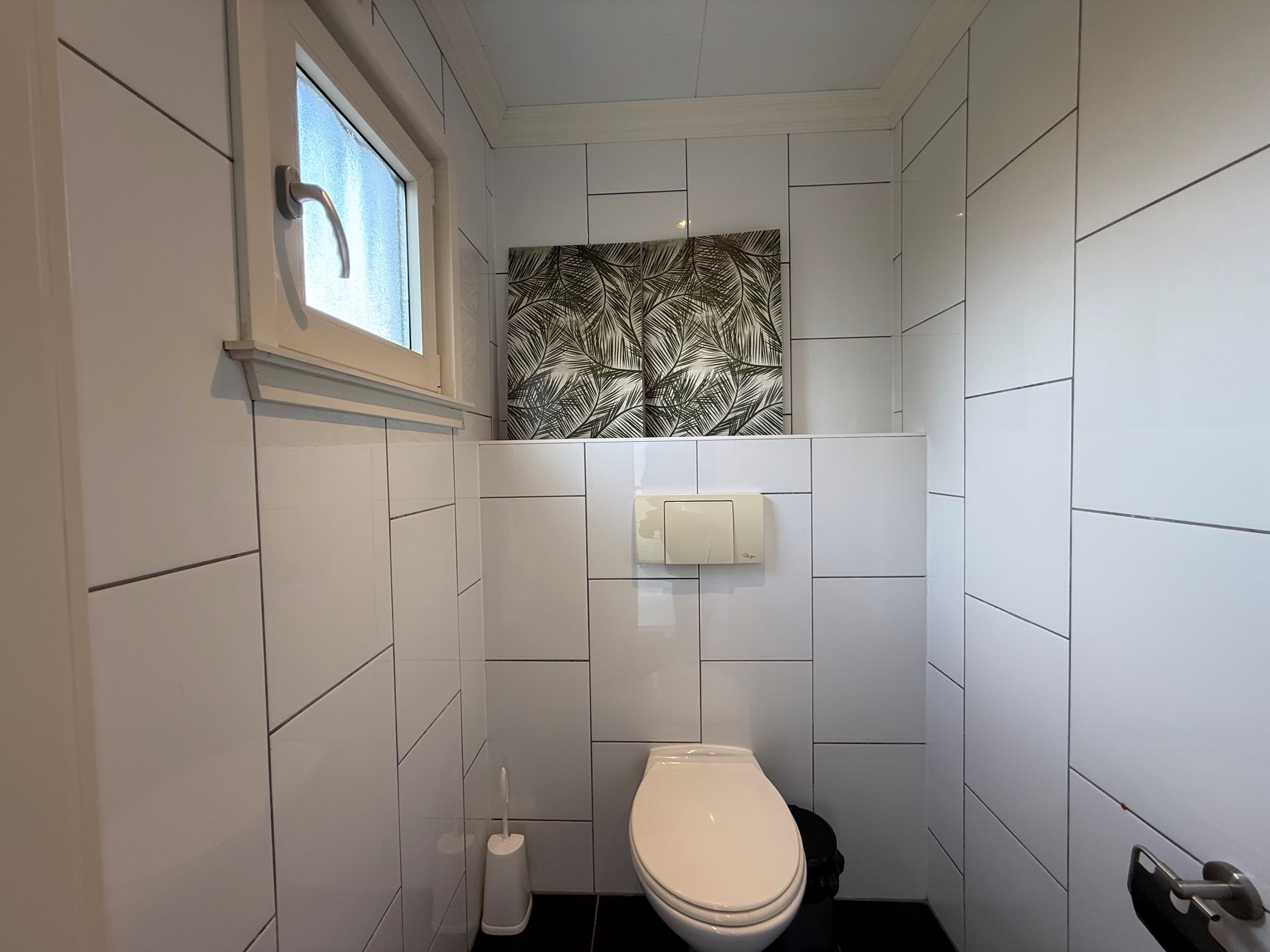 Klein, modern toilet met witte tegels en een plantenillustratie aan de muur.