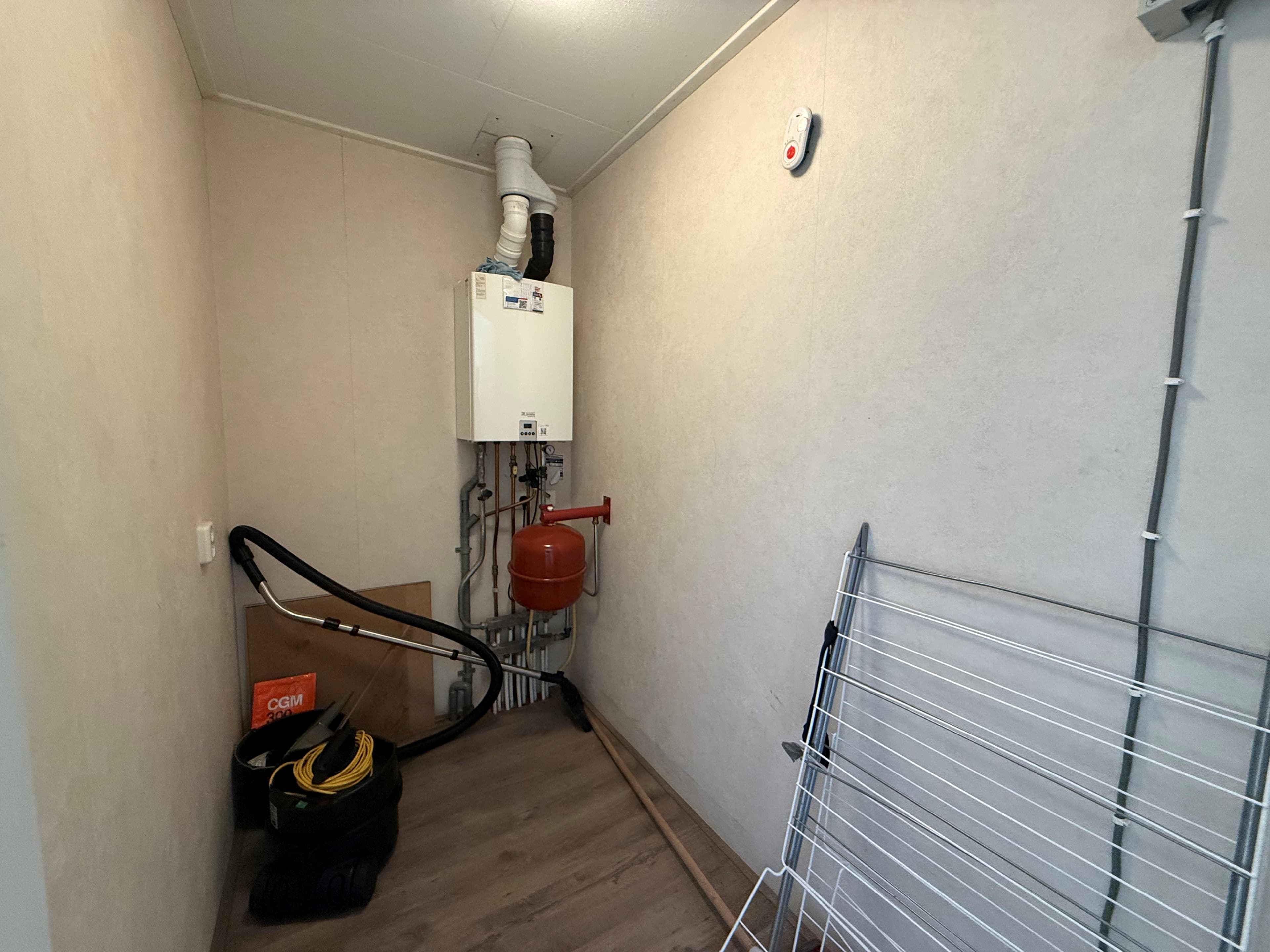 Kleine bijkeuken met een verwarmingsunit, waterleiding en ruimte voor opslag, inclusief een wasrek en een stofzuiger.