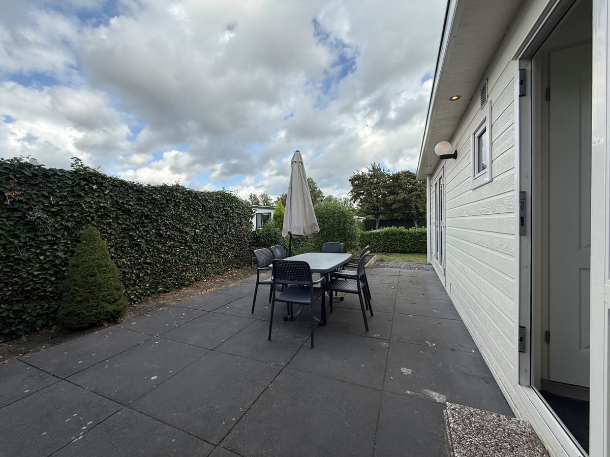 Terras met eettafel en stoelen bij een recreatiewoning, omringd door groene hagen en een blauwe lucht met wolken