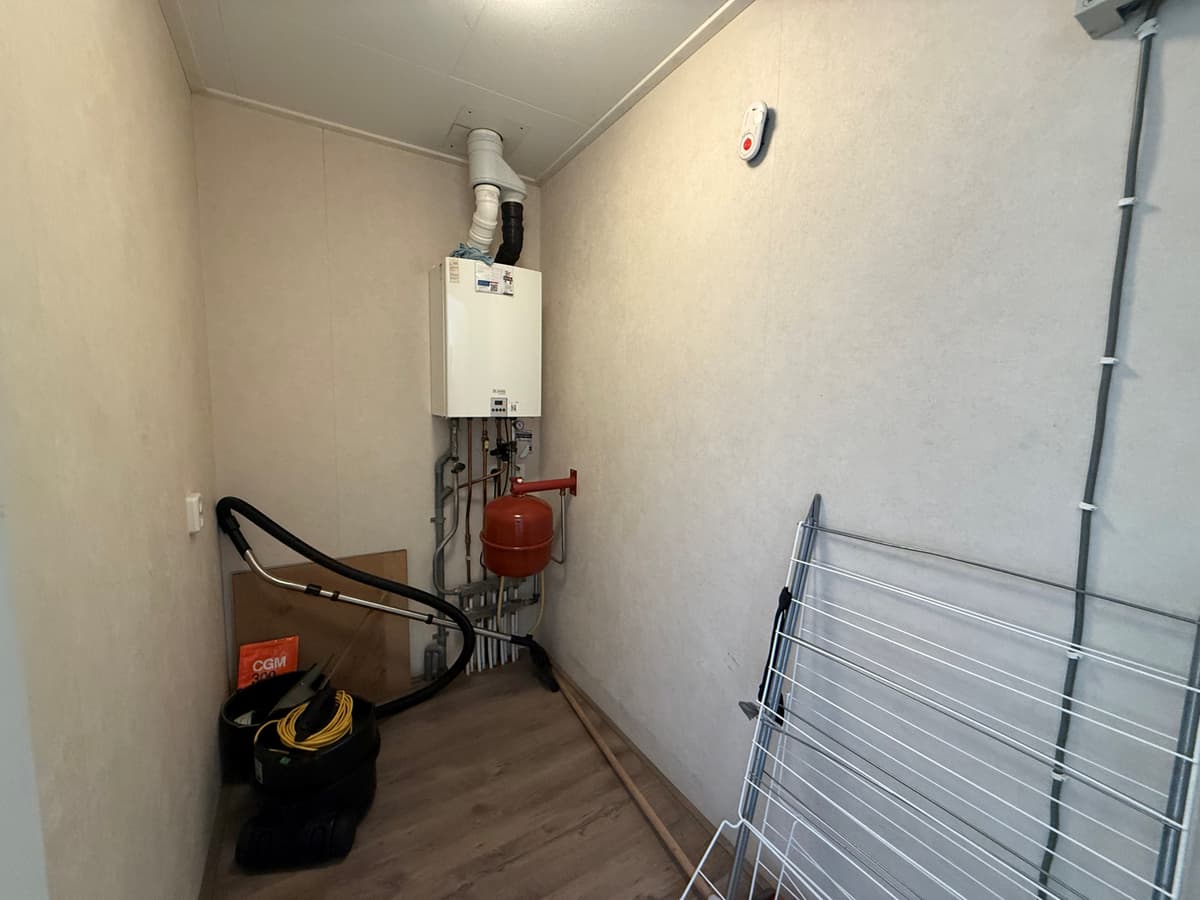 Kleine bijkeuken met een verwarmingsunit, waterleiding en ruimte voor opslag, inclusief een wasrek en een stofzuiger.