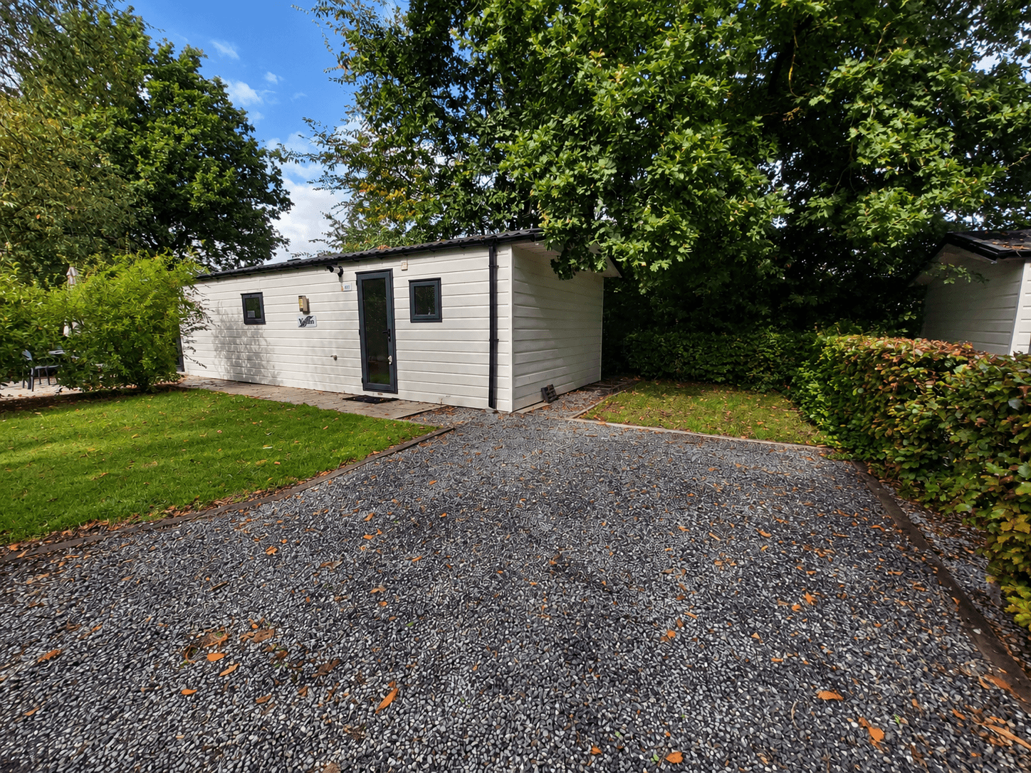 Witte recreatiewoning met zwarte deur en ramen, omringd door groen gras en bomen, uitzicht op grindpad.