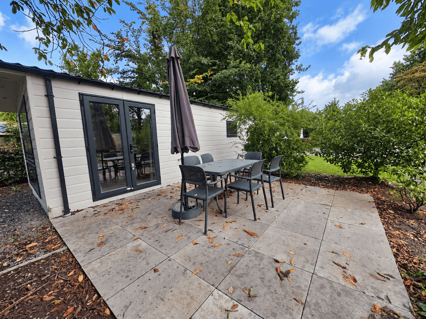 Terras met tuinmeubilair en parasol, grijze tegels, omliggend groen en blauwe lucht met enkele wolken.