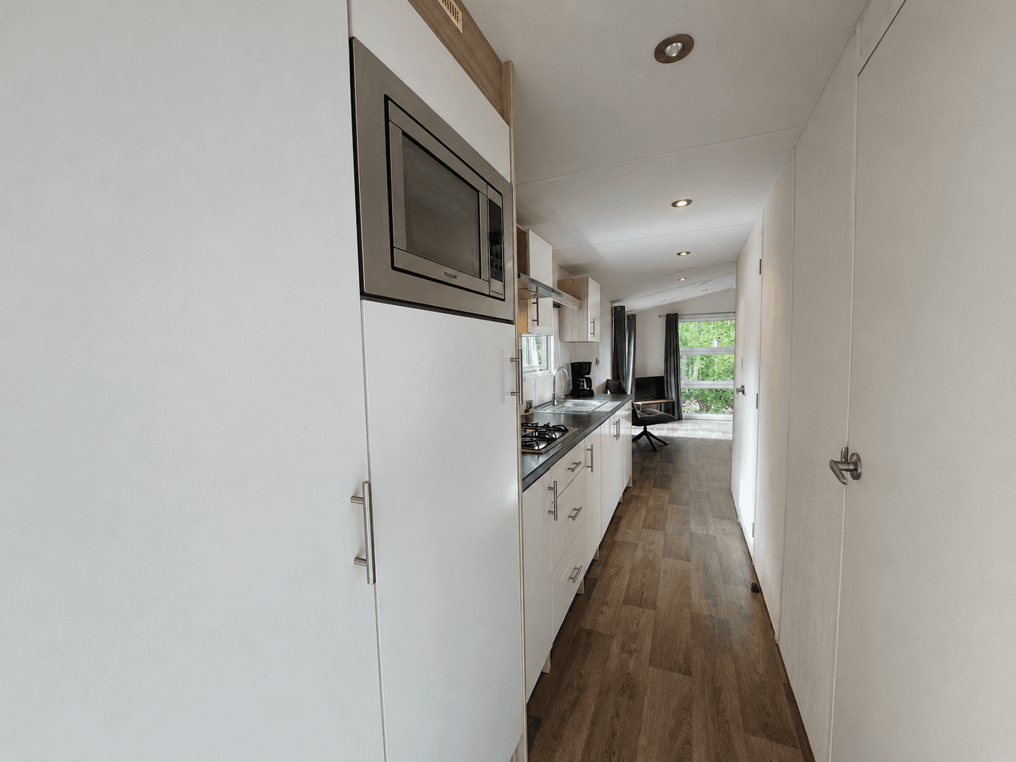 Keuken met inbouwapparatuur, gladde witte kasten en houten vloer, lichtinval van een raam aan het einde van de gang