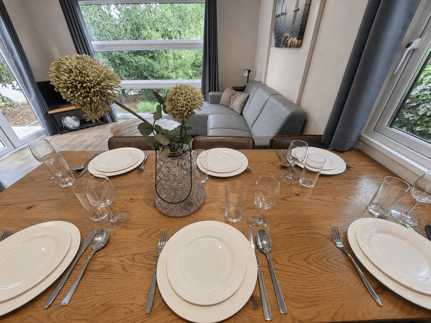 Eetkamertafel met wit servies, glazen en bestek, naast een grijze bank en grote ramen met uitzicht op groen buiten.