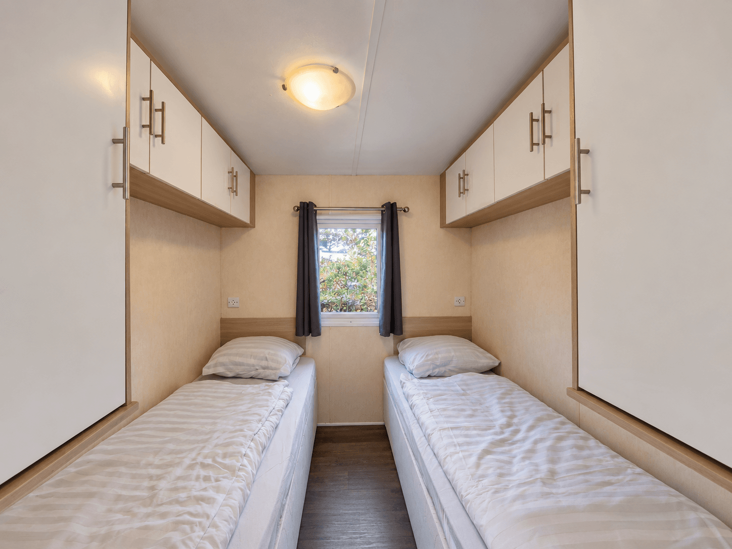 Slaapkamer met twee eenpersoonsbedden, bovenkasten en een raam met zicht op groen.