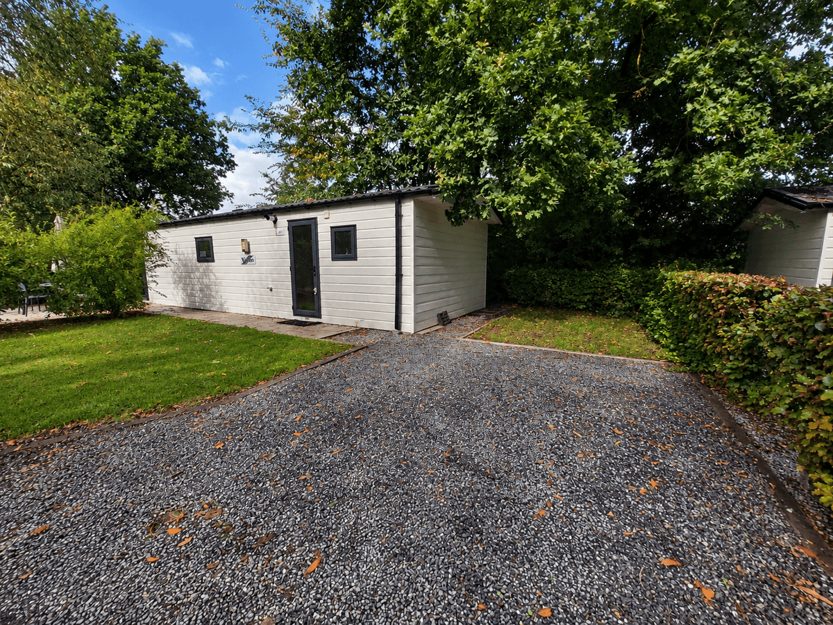 Witte recreatiewoning met zwarte deur en ramen, omringd door groen gras en bomen, uitzicht op grindpad.