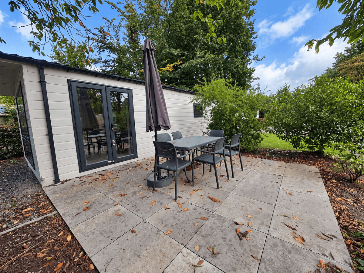 Terras met tuinmeubilair en parasol, grijze tegels, omliggend groen en blauwe lucht met enkele wolken.
