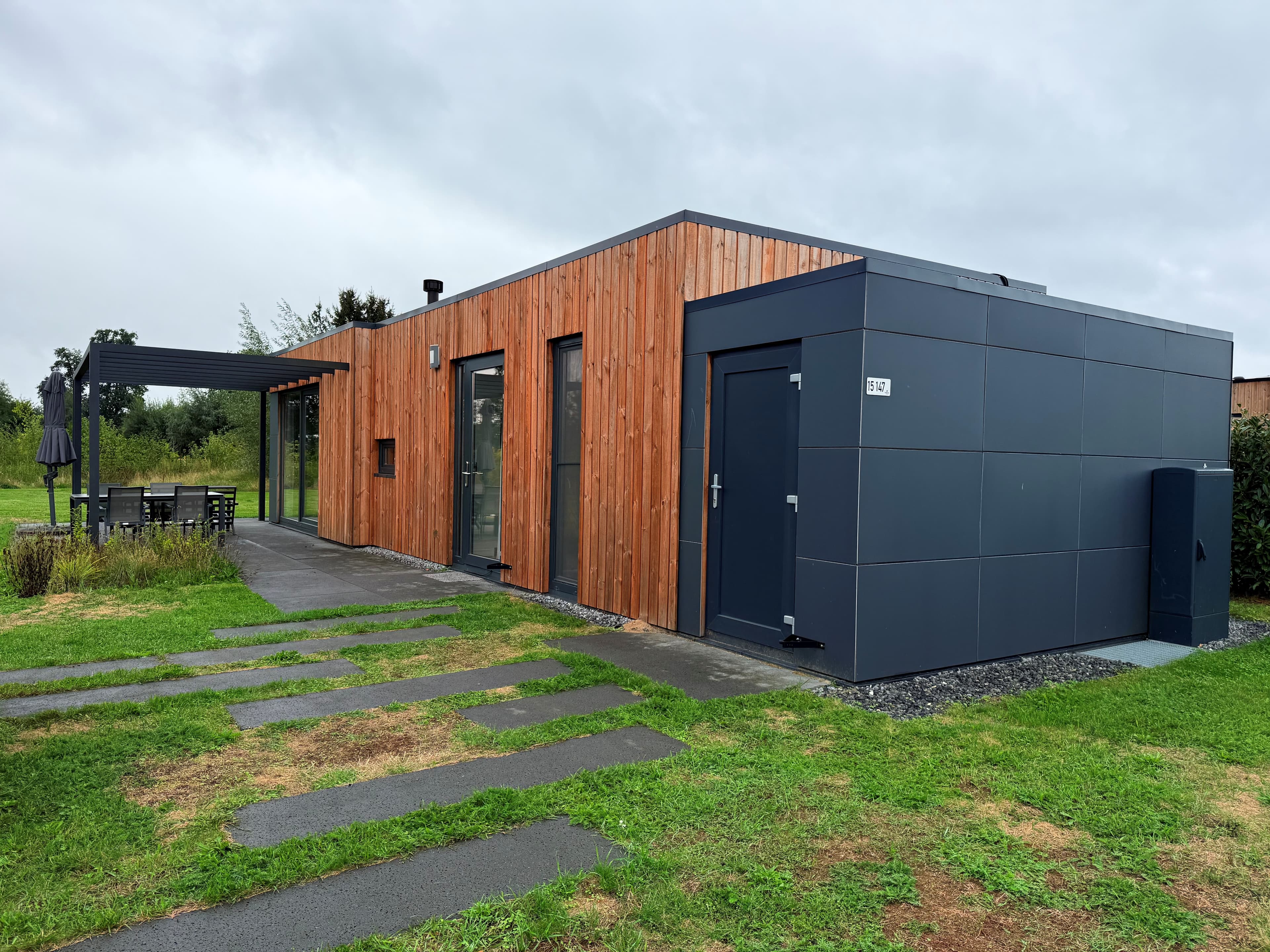 Recreatiewoning met houten gevel en moderne aanbouw, omlijst door gras en bestrating, gelegen op vakantiepark.