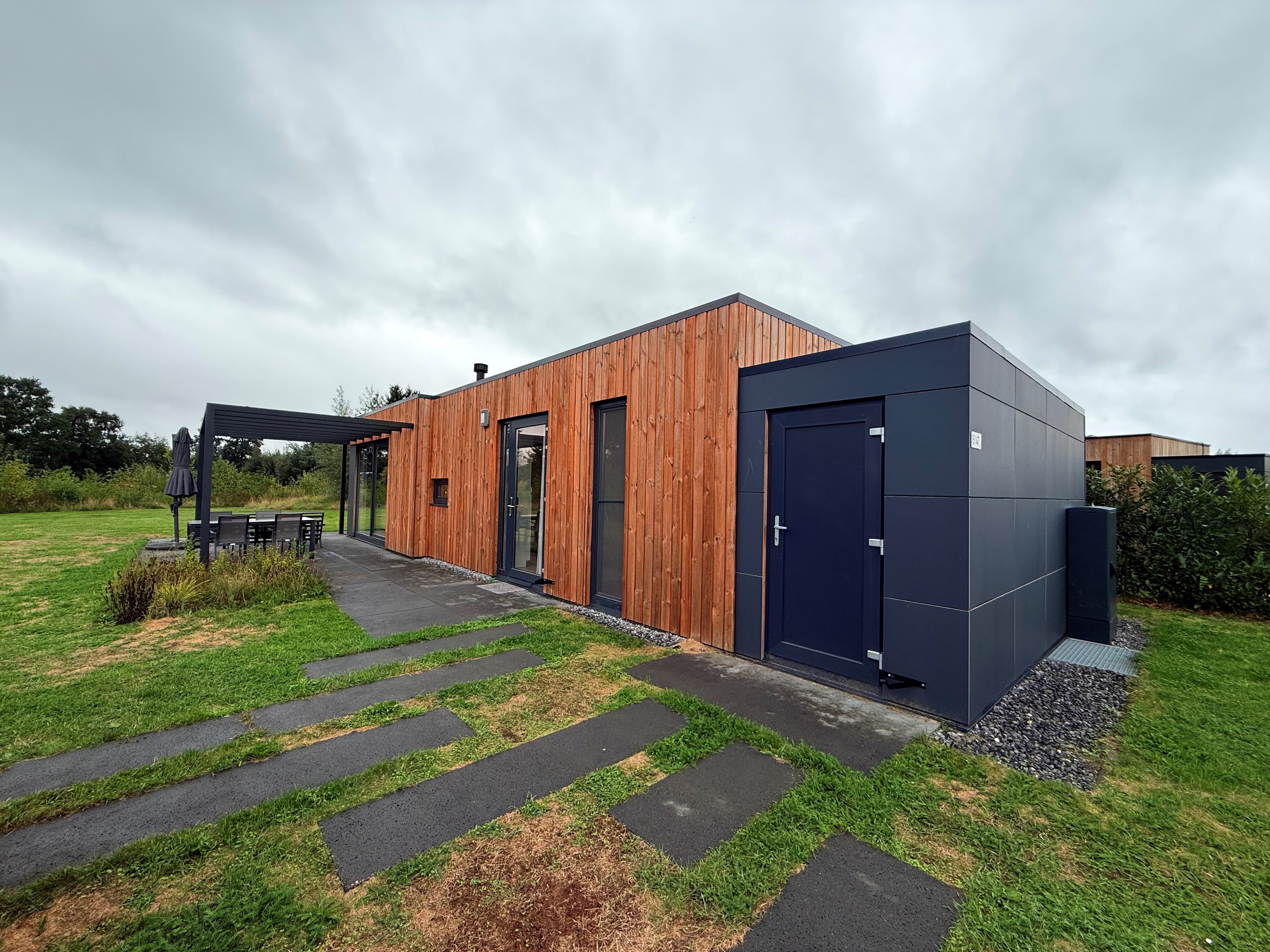 Moderne recreatiewoning met houten gevel en antracietkleurige accenten, gelegen op een groen vakantiepark met betegelde paden