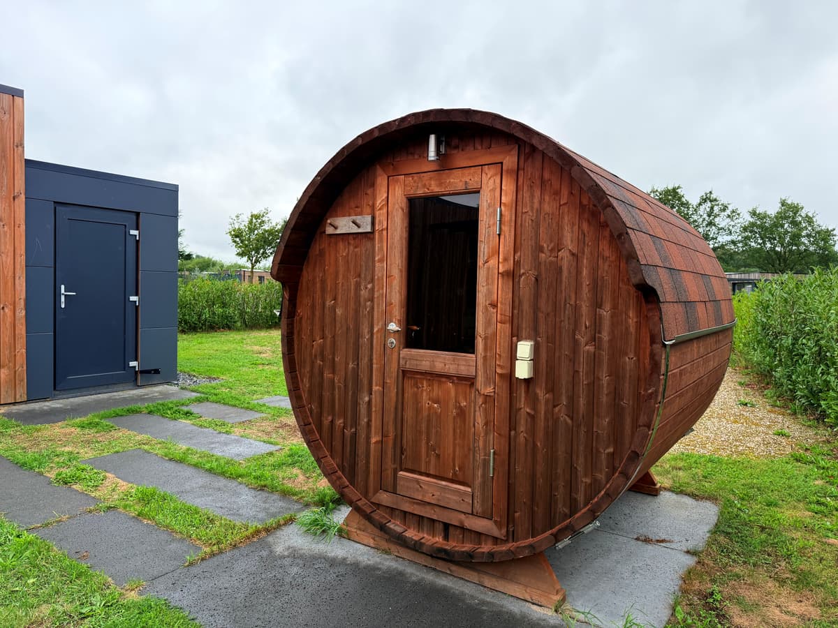 Houten barrelvormige sauna met deur, gelegen op een gazon bij een vakantiehuis in een groene omgeving