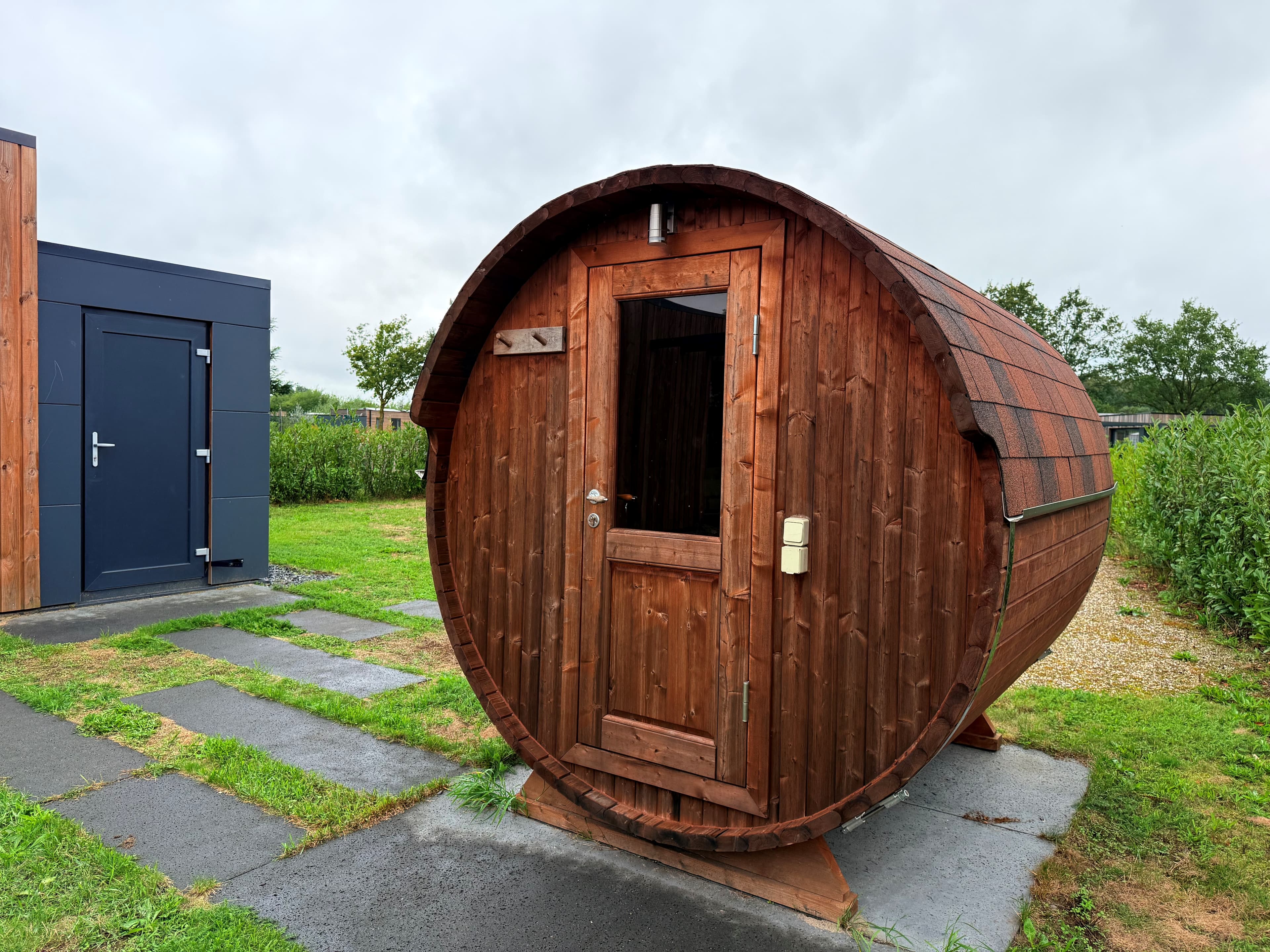 Houten barrelvormige sauna met deur, gelegen op een gazon bij een vakantiehuis in een groene omgeving
