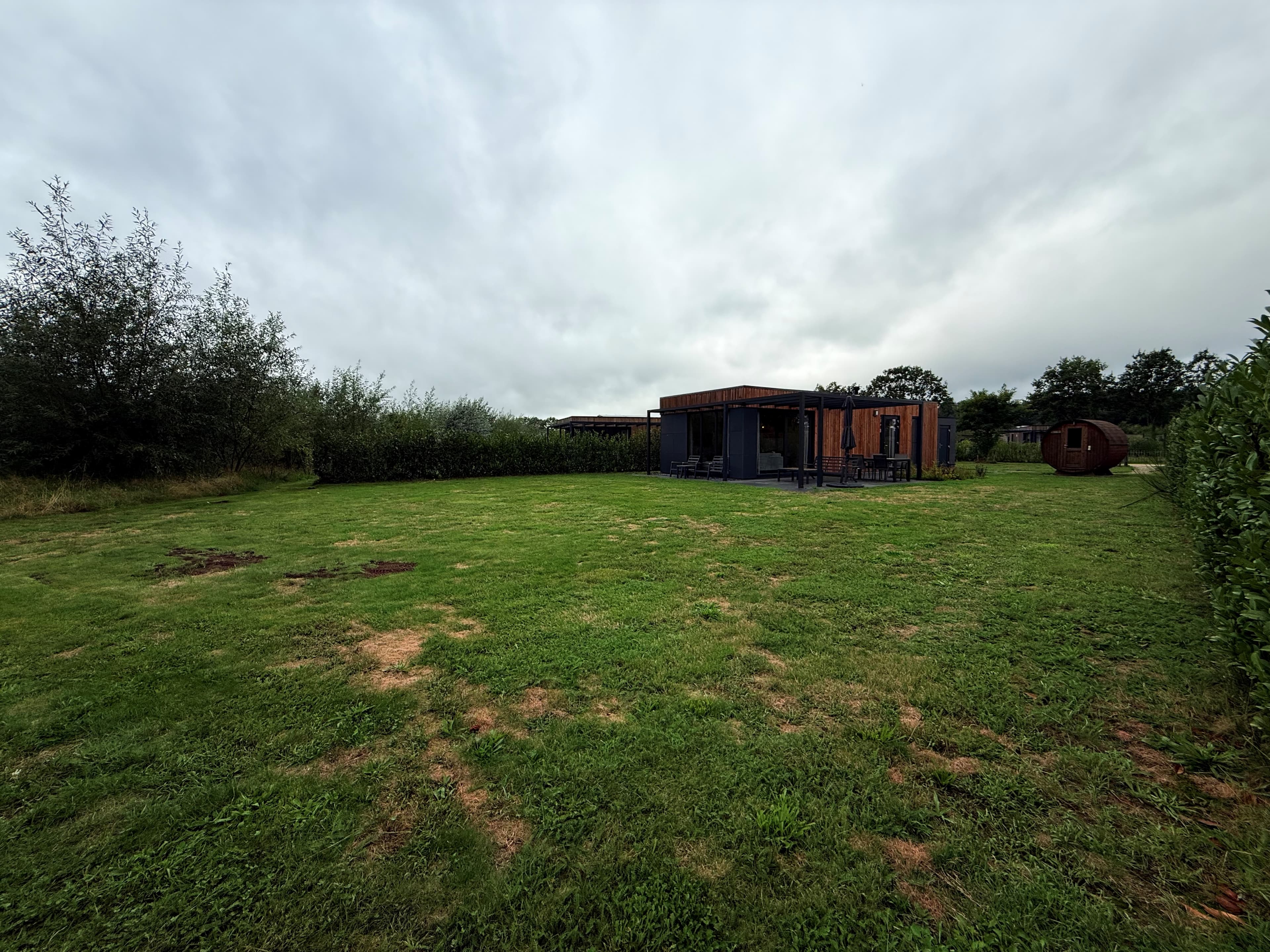 Recreatiewoning op een grasveld omringd door bomen, met een terras en een houten schuur op de achtergrond.