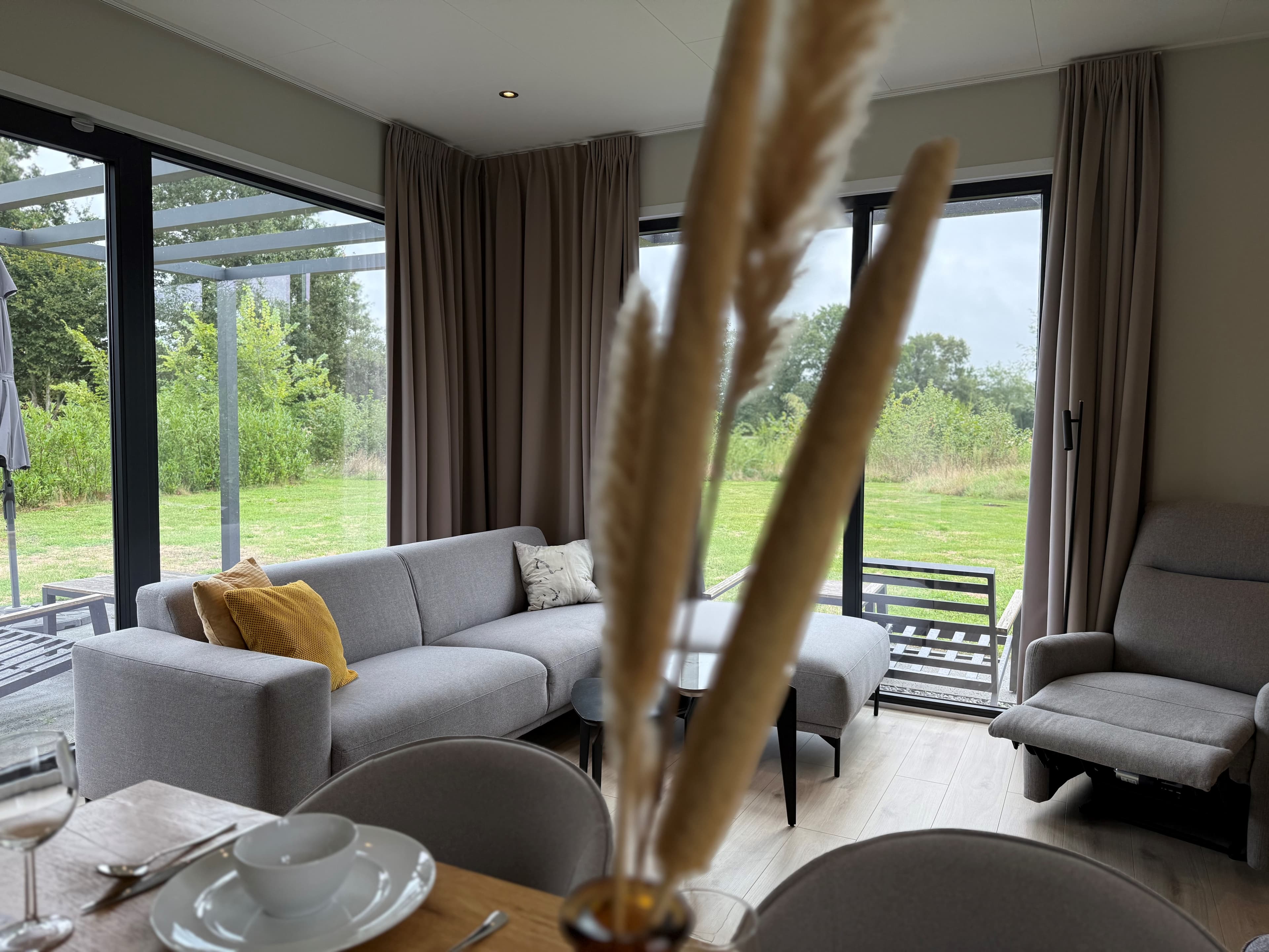 Lounge met zithoek en eettafel, grote ramen met uitzicht op de groene tuin en natuur buiten.