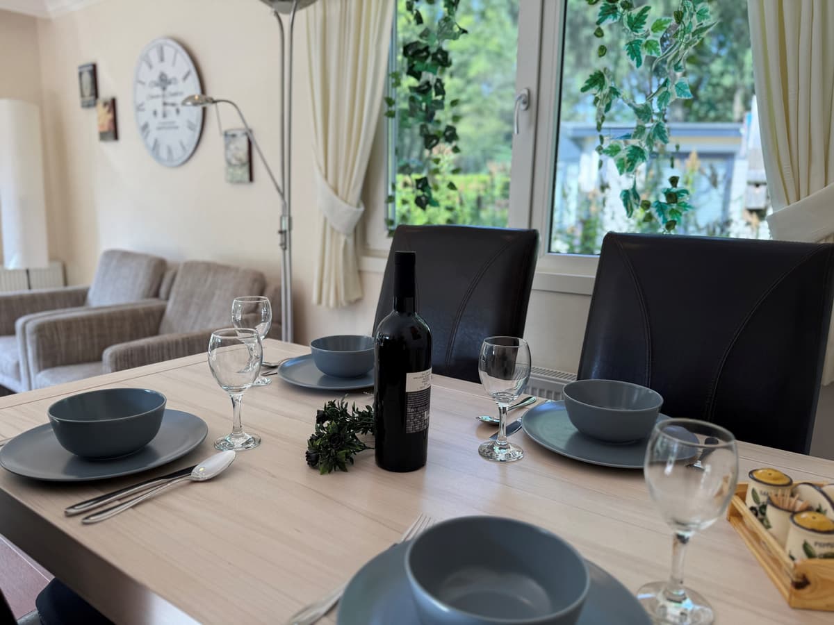 Eetkamer met een gedekte tafel, vier stoelen, een fles wijn en uitzicht op de tuin door het raam
