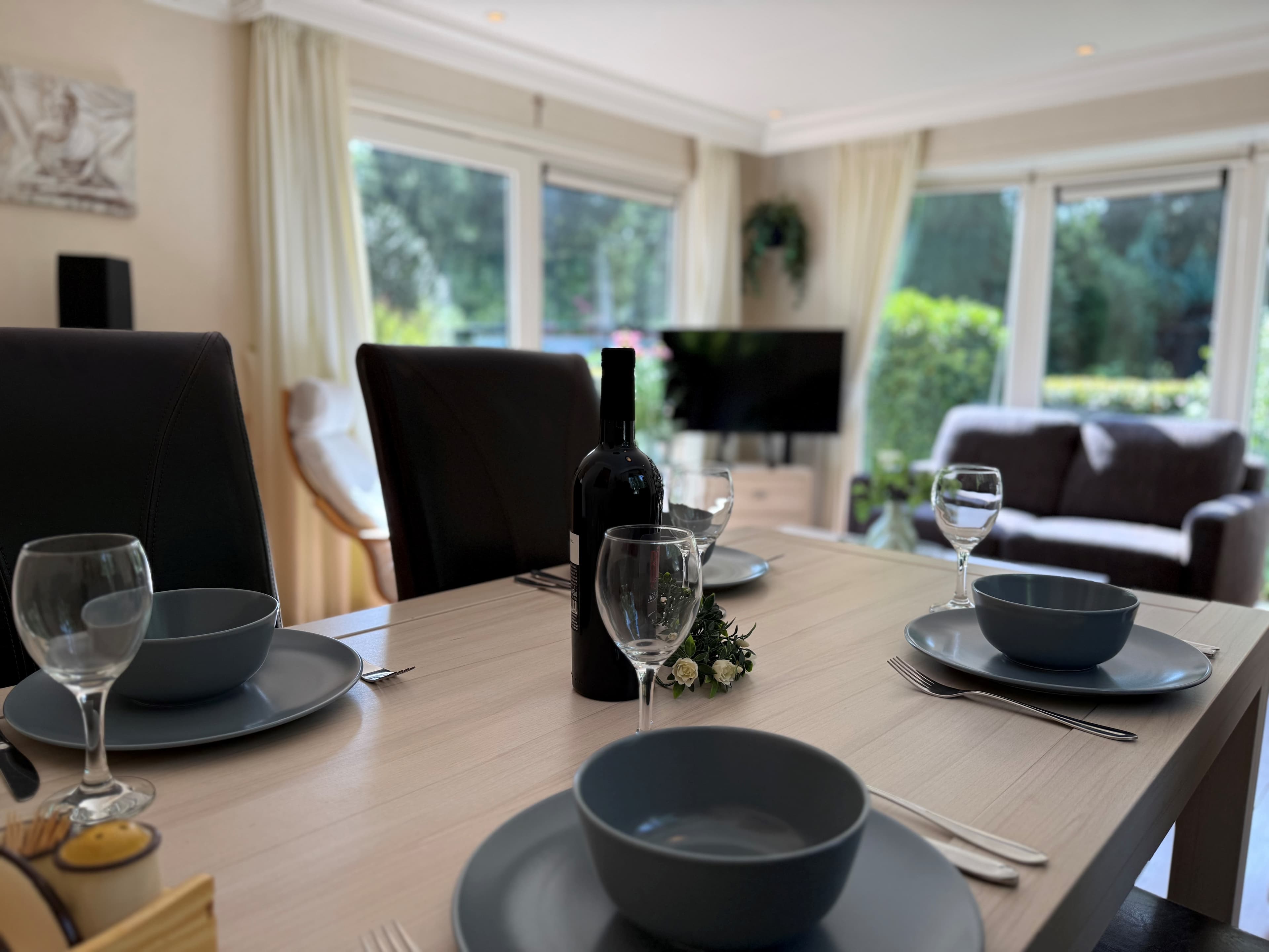 Eettafel met vier borden, glazen en een fles wijn, uitzicht op een woonkamer met bank en tv, veel natuurlijk licht