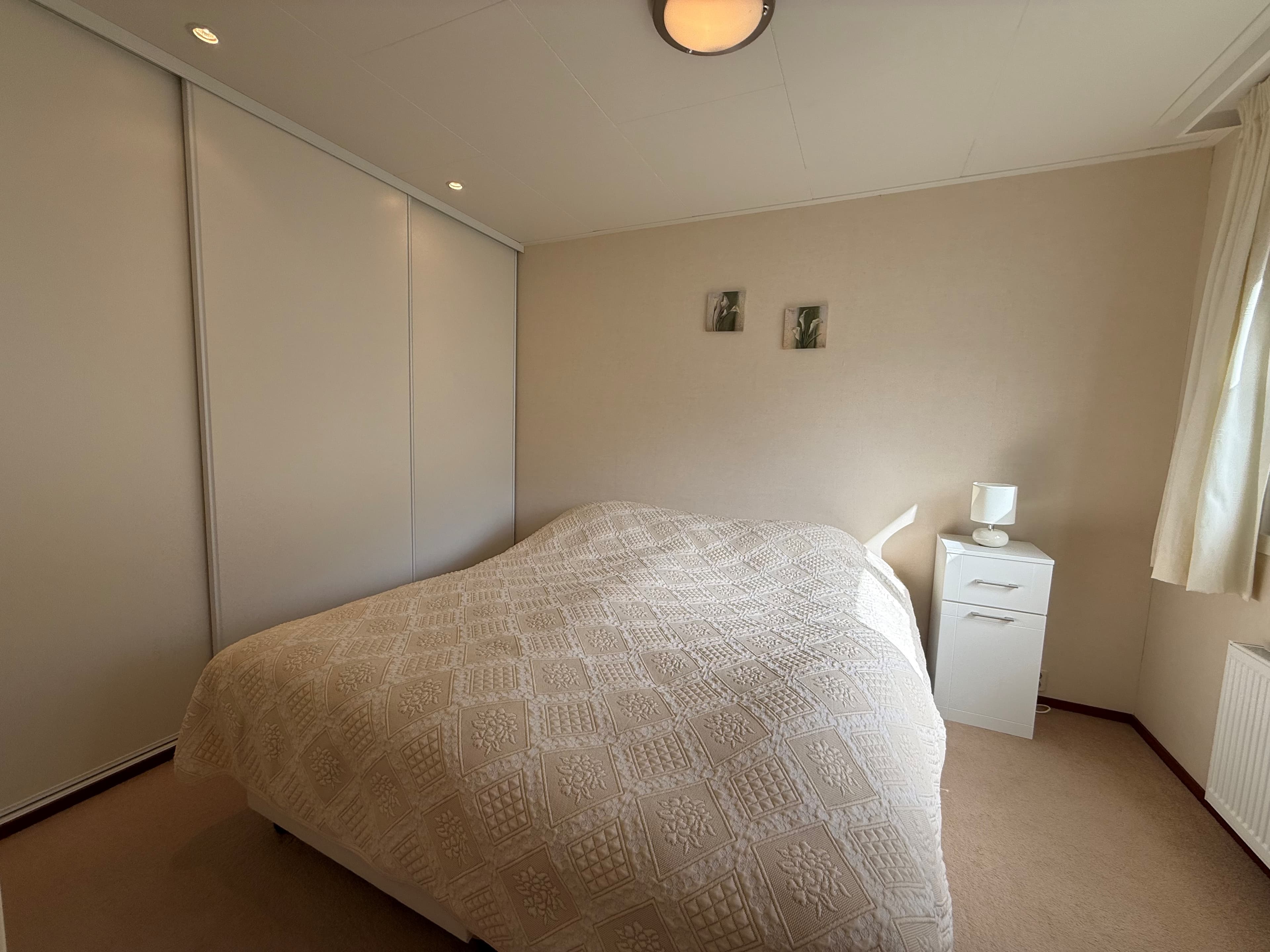 Slaapkamer met een tweepersoonsbed, witte kast en nachtkastje, voorzien van een raam met gordijnen.