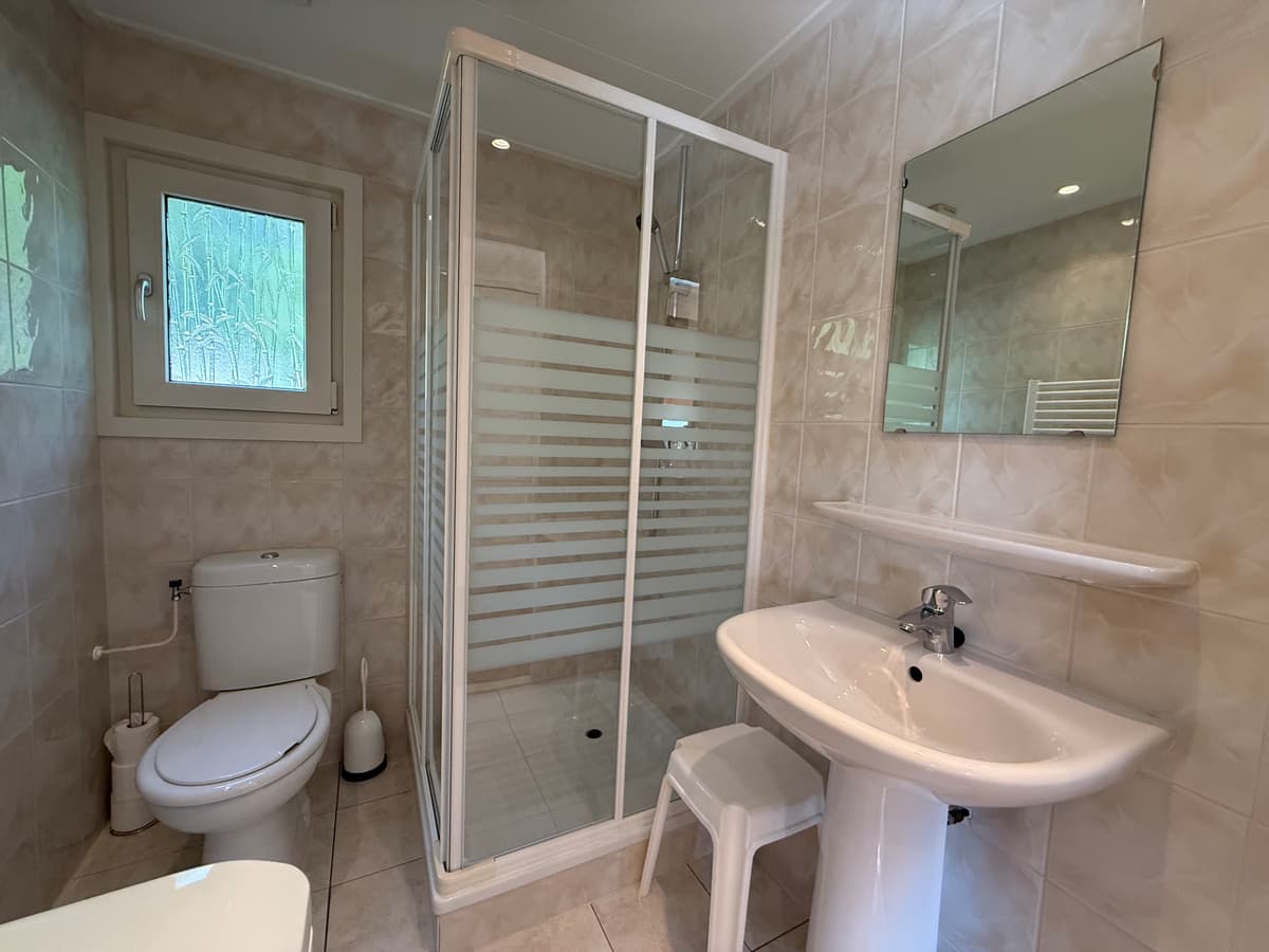Badkamer met douchecabine, wastafel en toilet in lichte tegelkleur met een raam voor natuurlijk licht.