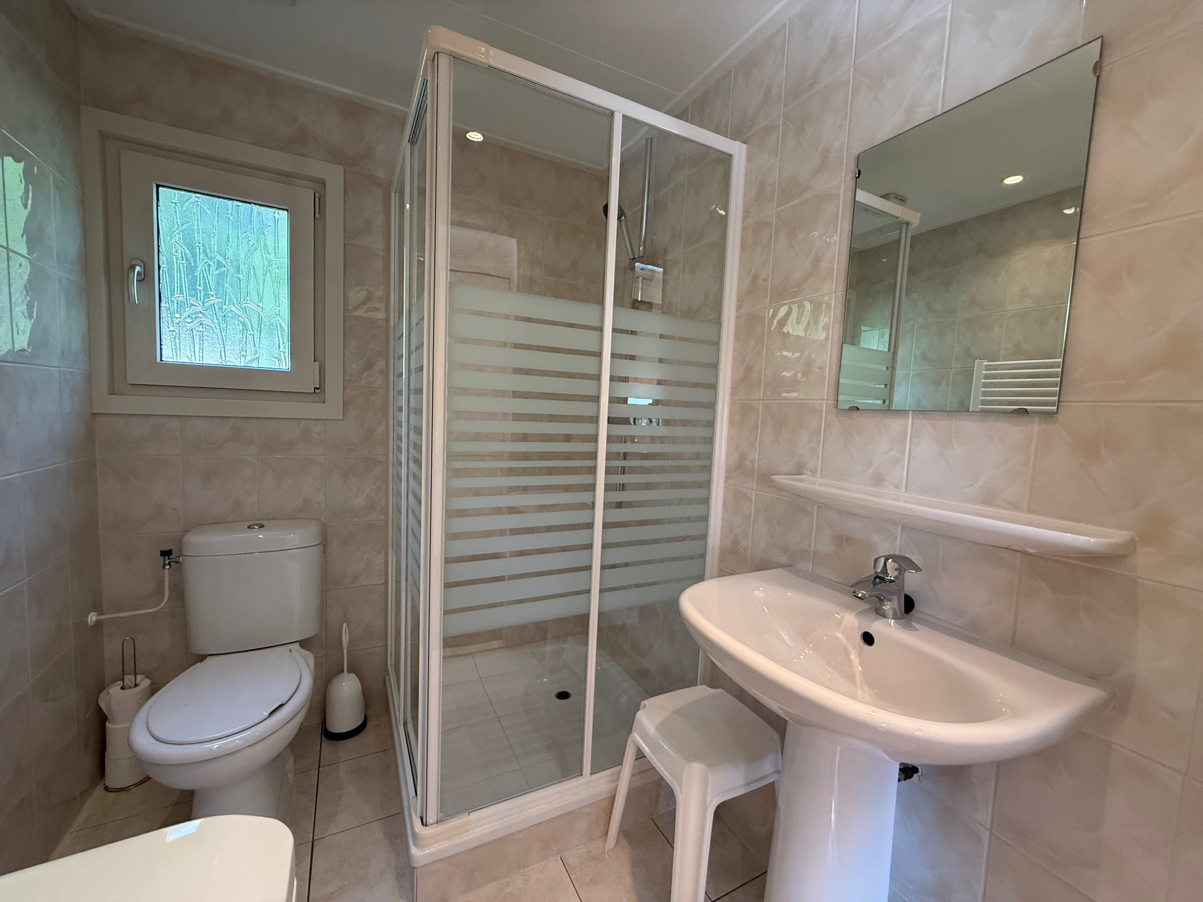 Badkamer met douchecabine, wastafel en toilet in lichte tegelkleur met een raam voor natuurlijk licht.