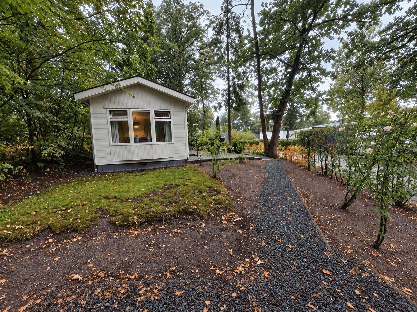 Witte recreatiewoning omringd door bomen en een betegeld pad met rozenstruiken en gras.