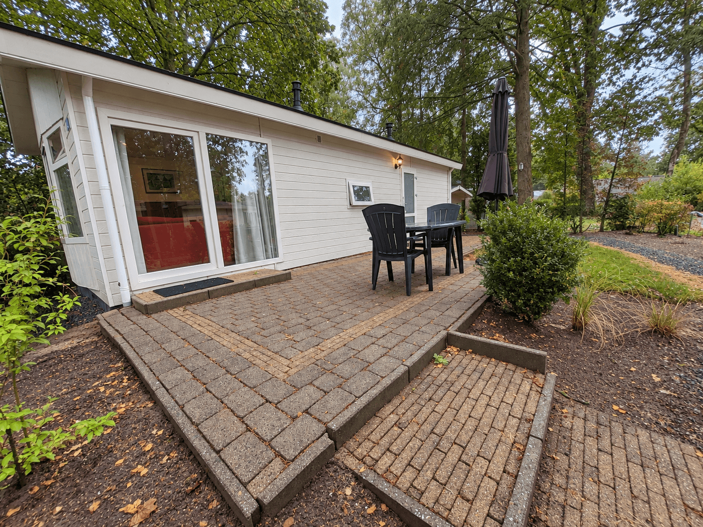 Zijaanzicht van een recreatiewoning met een terras van stenen, een parasol en groene planten eromheen