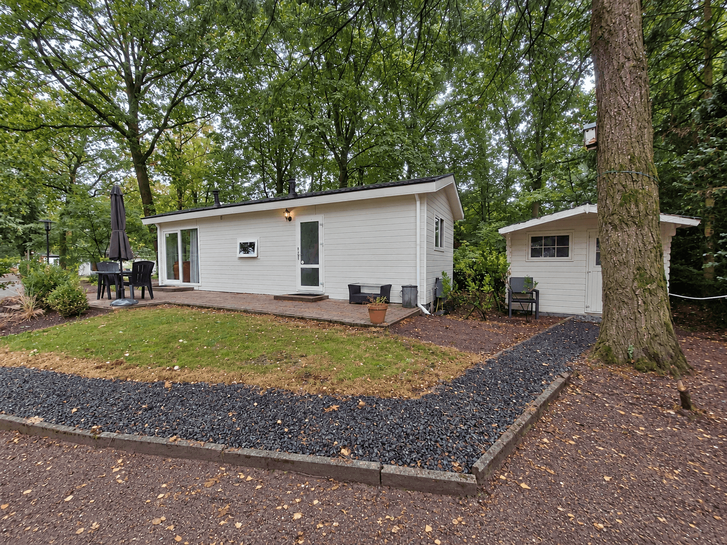 Recreatiewoning met wit houten gevel, een terras met stoelen en een naastgelegen tuinhuisje omgeven door bomen