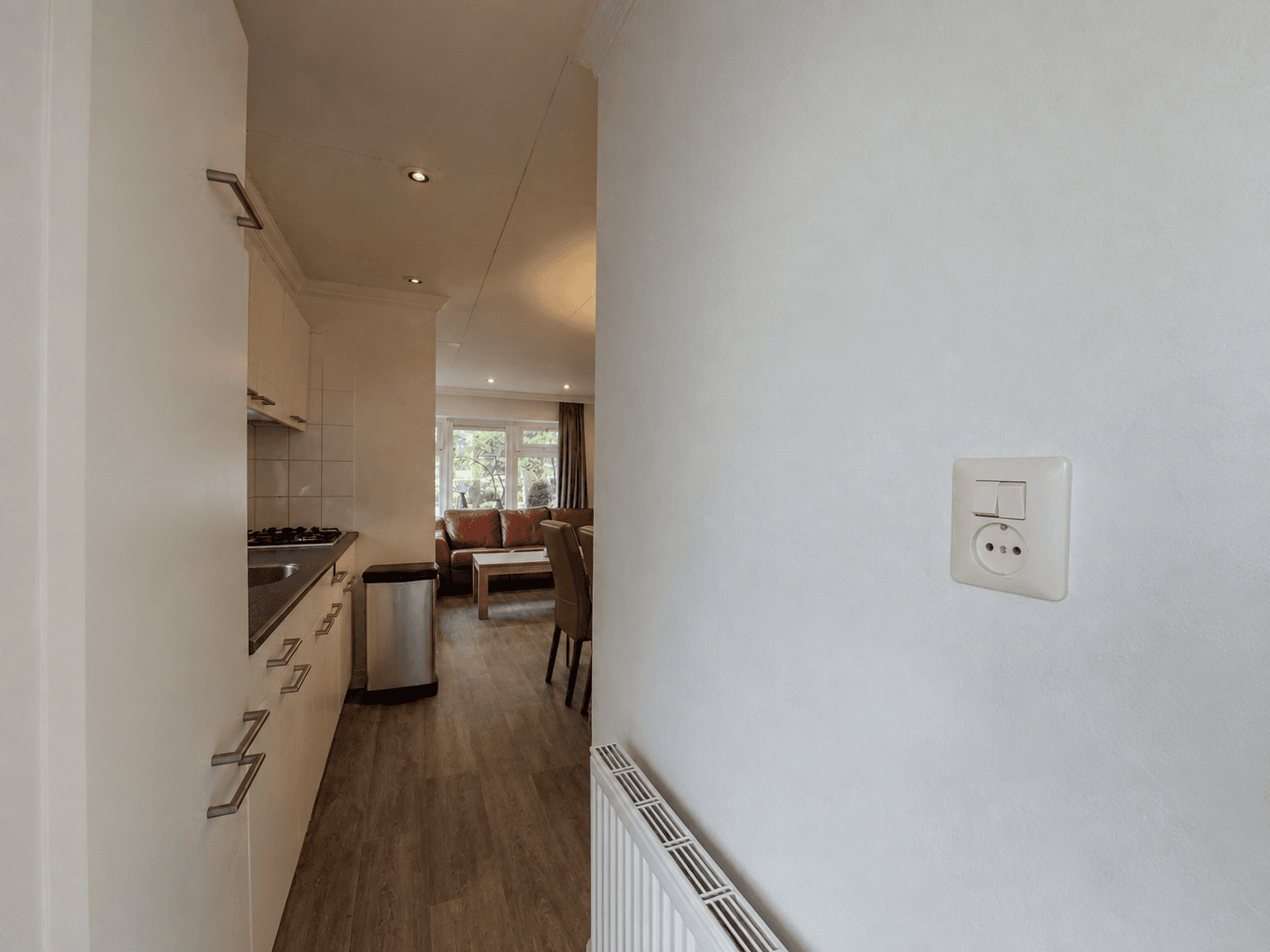 Zicht op de keuken met lichte kastjes en een open gang naar de woonkamer met een bank en eettafel