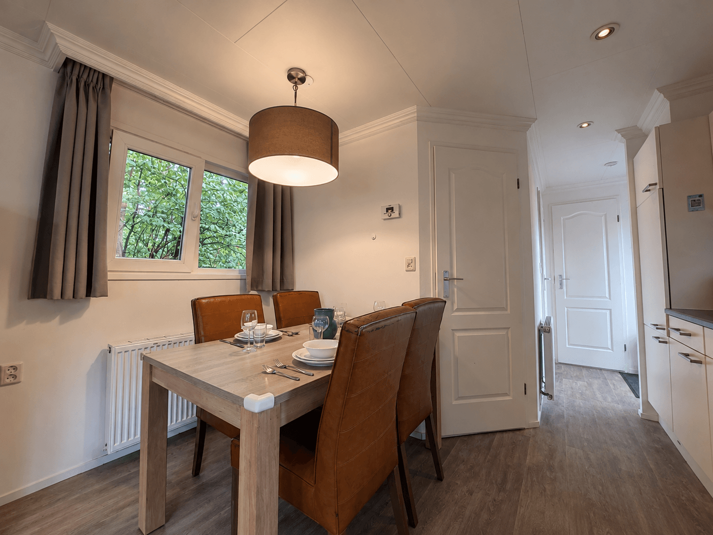 Eetruimte met een houten tafel en bruine stoelen, ramen met zicht op groen, lichte muren en moderne verlichting