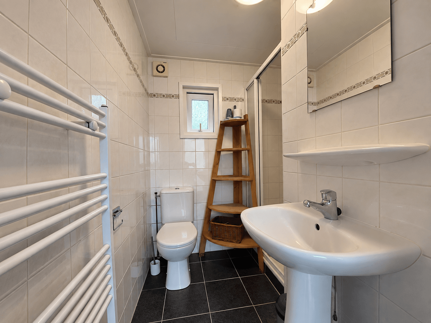 Badkamer met toilet, wastafel, spiegel en doucheschuifwand; wit betegelde wanden en een houten ladder voor opbergruimte