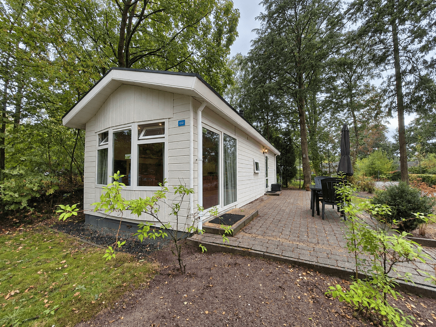Witte recreatiewoning met terras, omringd door groen en bomen, gelegen in een rustige omgeving met buitenmeubilair.