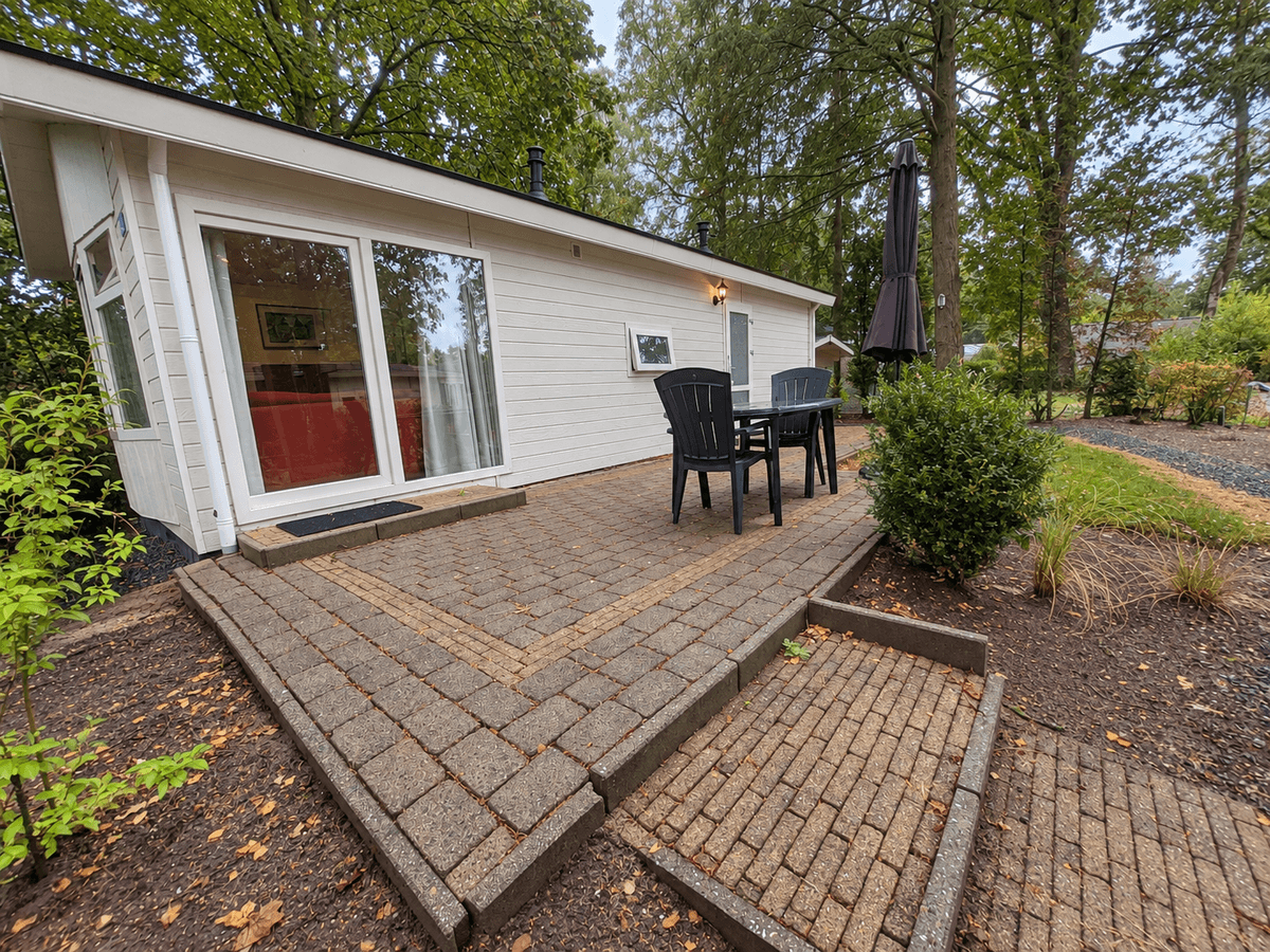 Zijaanzicht van een recreatiewoning met een terras van stenen, een parasol en groene planten eromheen