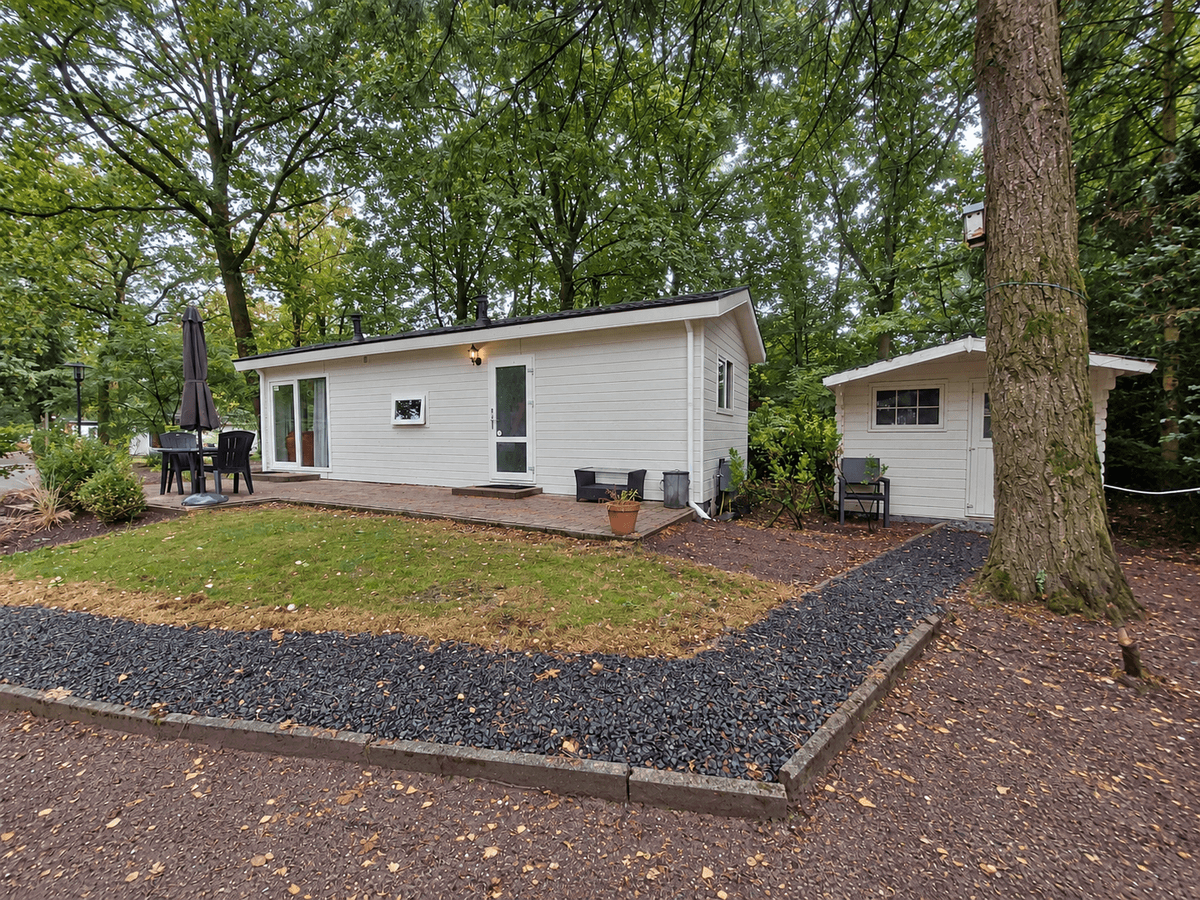 Recreatiewoning met wit houten gevel, een terras met stoelen en een naastgelegen tuinhuisje omgeven door bomen