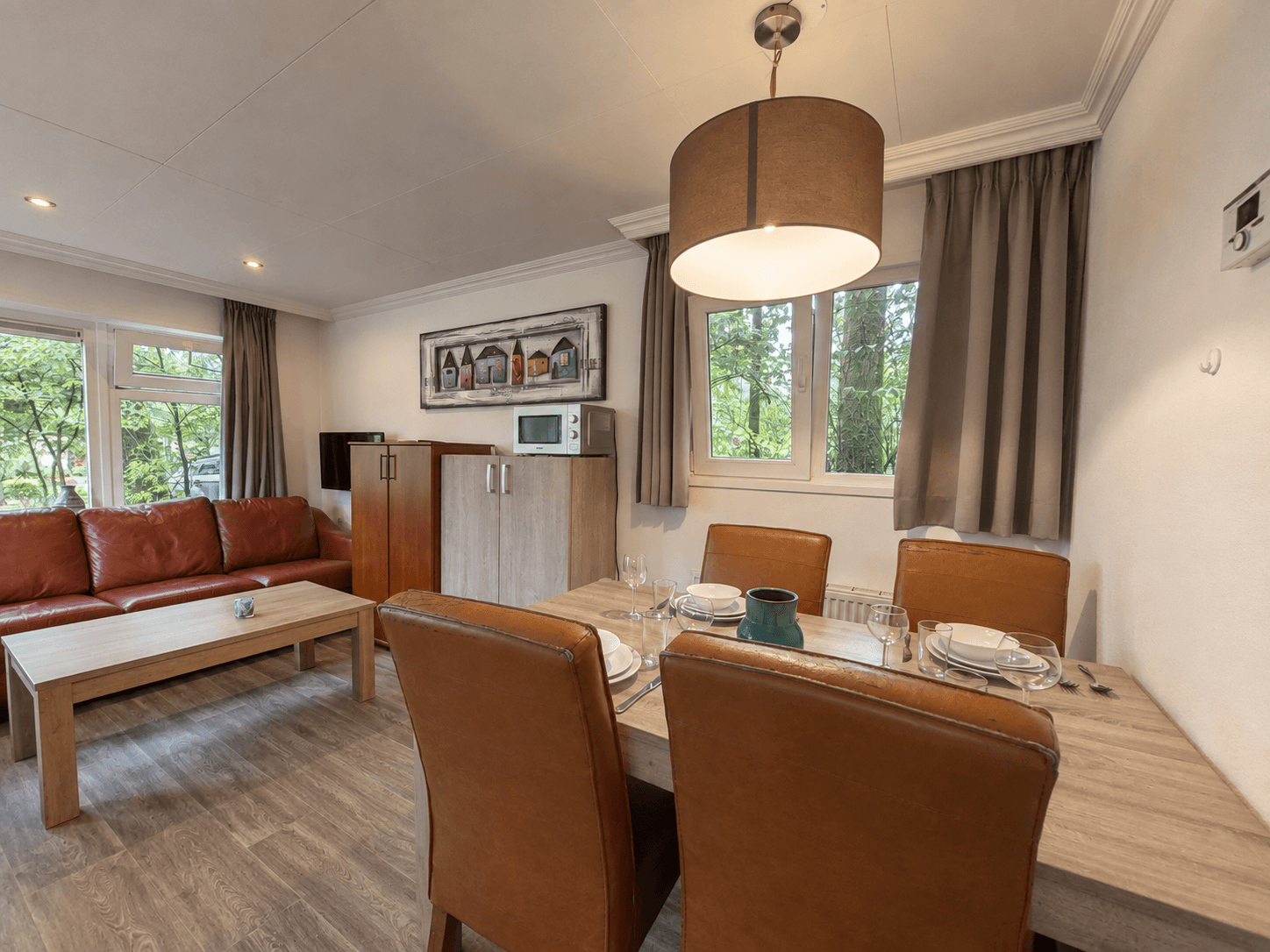 Eet- en woonruimte met een houten tafel, stoelen, bank, keukenhoek en ramen met uitzicht op bomen.