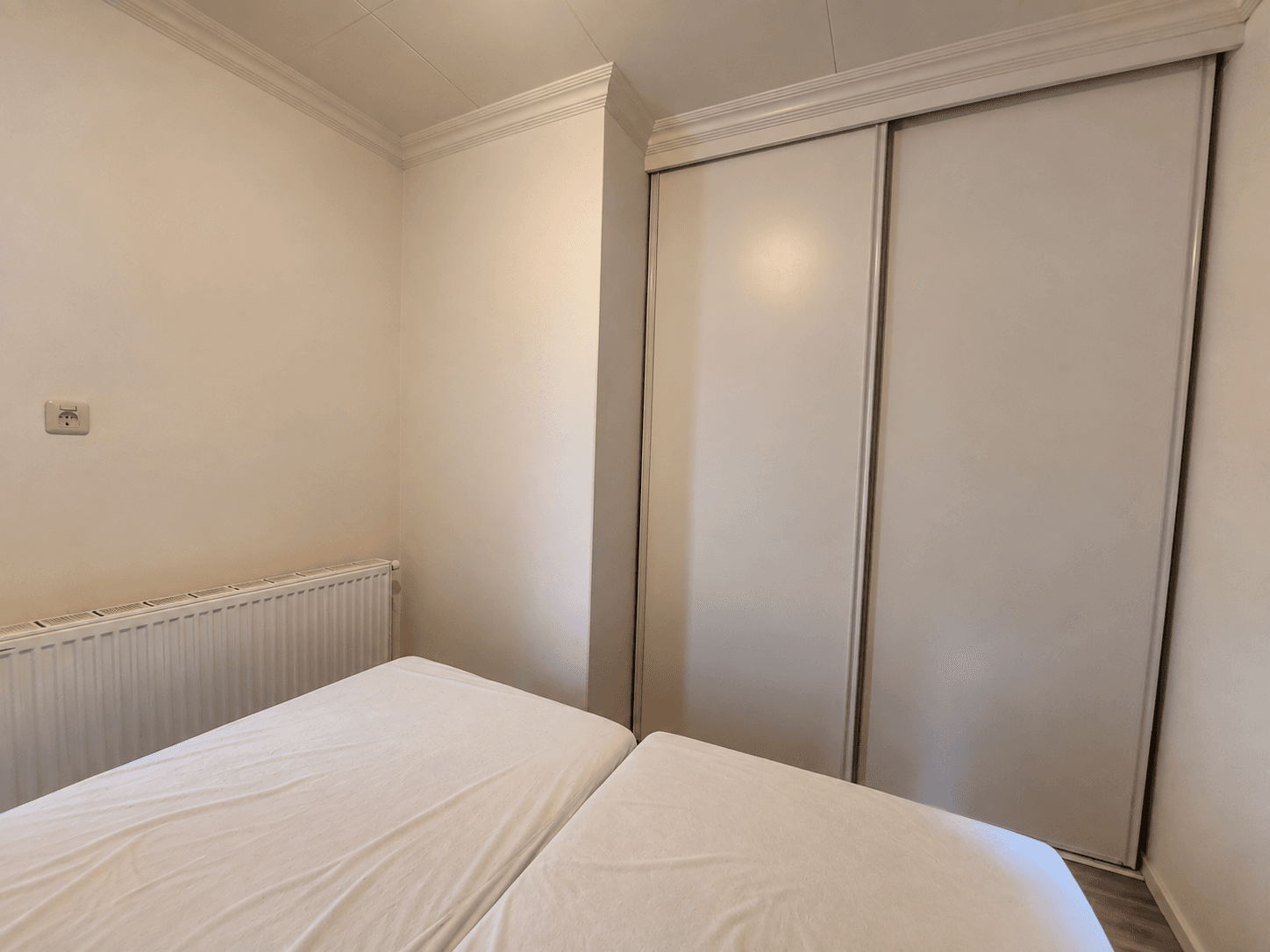 Slaapkamer met twee bedden en een inbouwkast, lichte wandafwerking en radiator zichtbaar