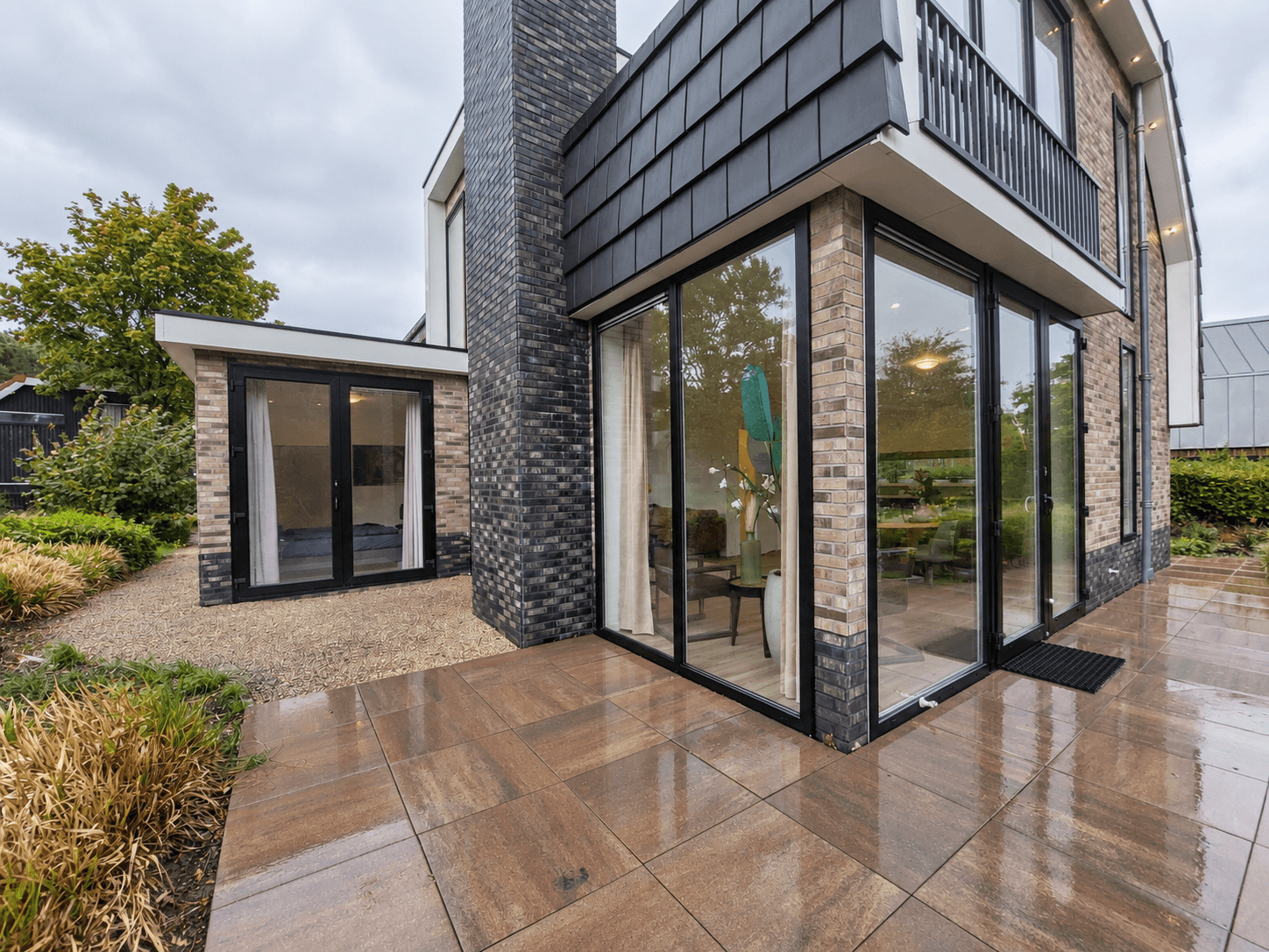 Moderne recreatiewoning met grote ramen, een tegelterras en een tuin met diverse planten en een grasveld
