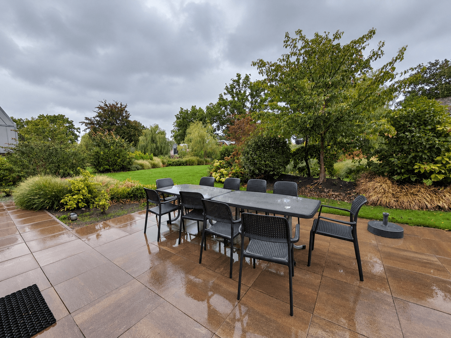 Tuin met een betegeld terras en een grote tafel met stoelen, omringd door groen en bomen onder een bewolkte lucht