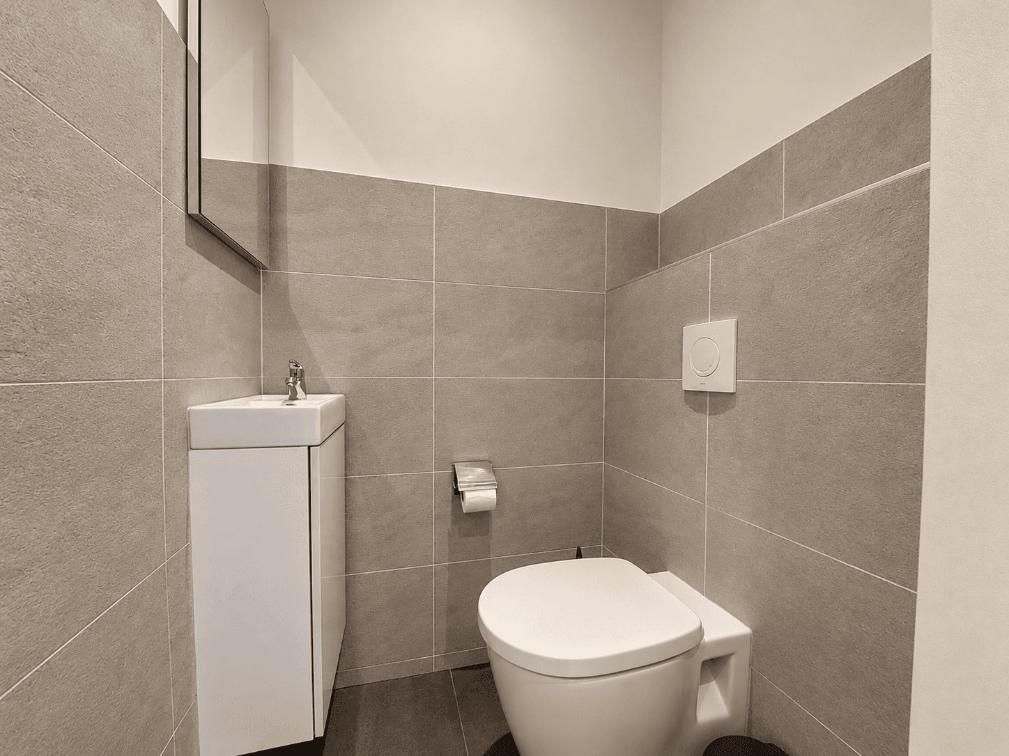 Modern toilet met beige en grijze tegels, een klein wastafeltje en een spiegel aan de muur.